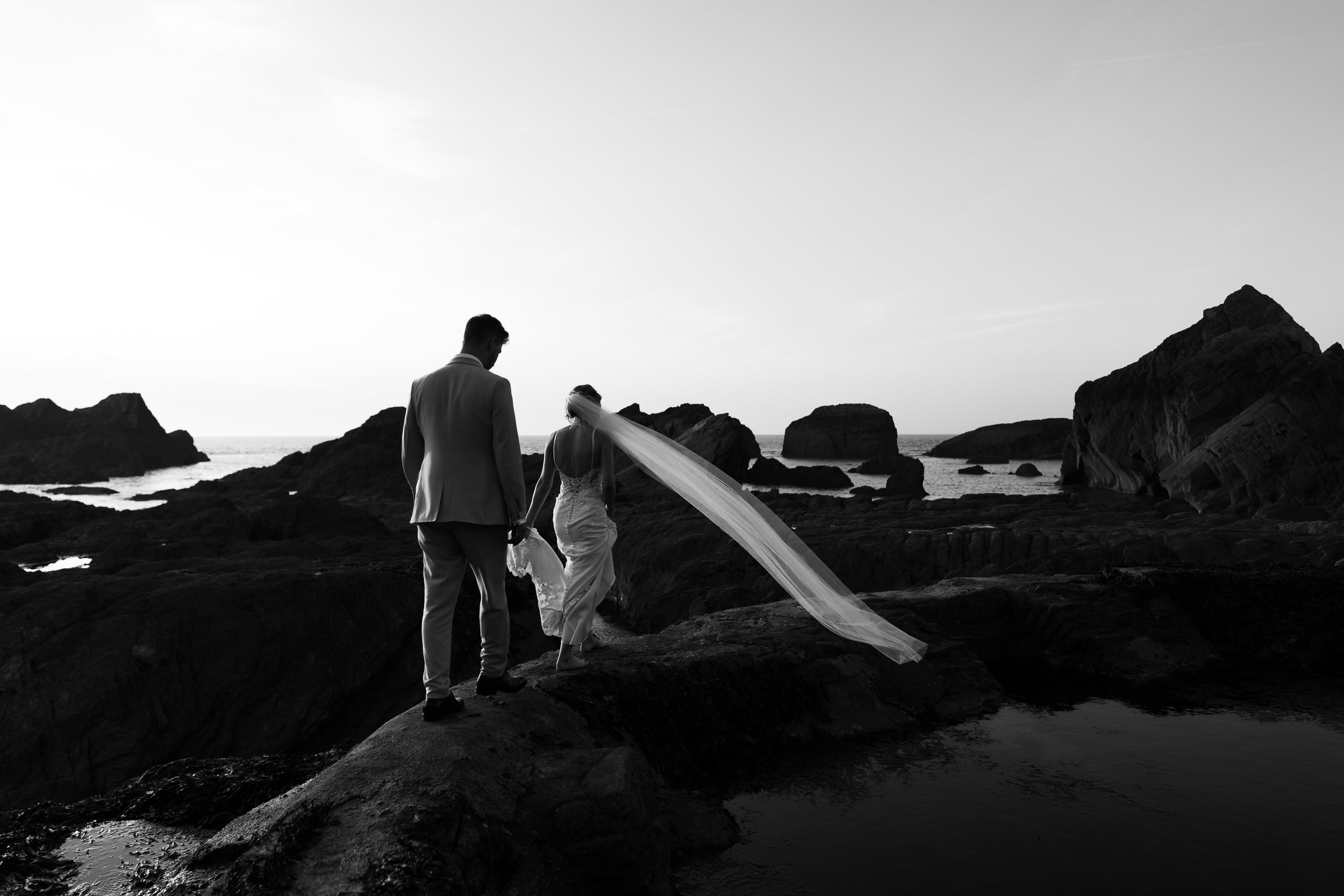 Bride and groom walking on rocky coast at sunset at Tunnels Beach, black and white photo.