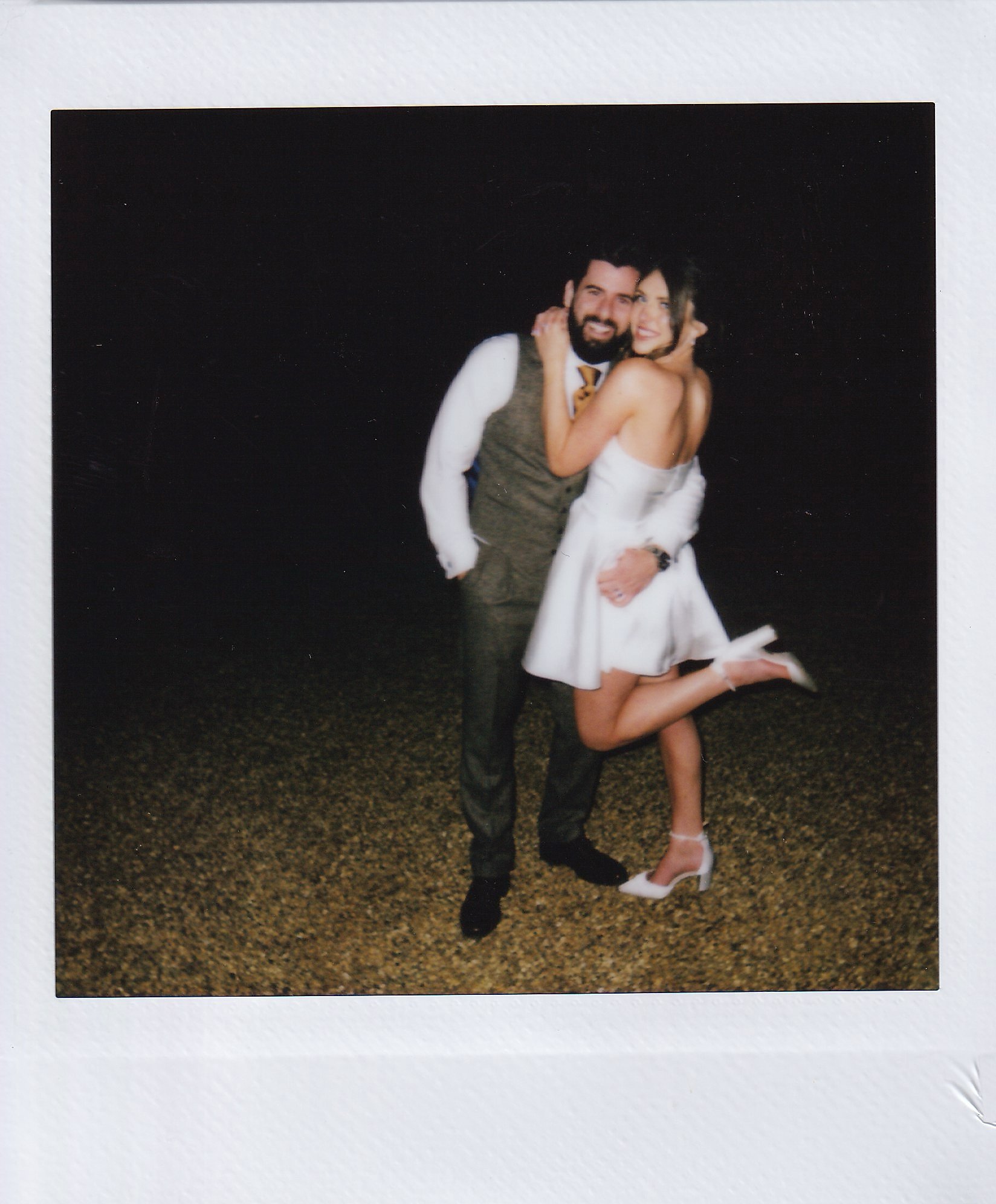 A couple posing happily at night. The man is wearing a suit with a waistcoat, and the woman is in a short white dress with heels. She is playfully lifting one leg. Both are smiling and embracing each other.