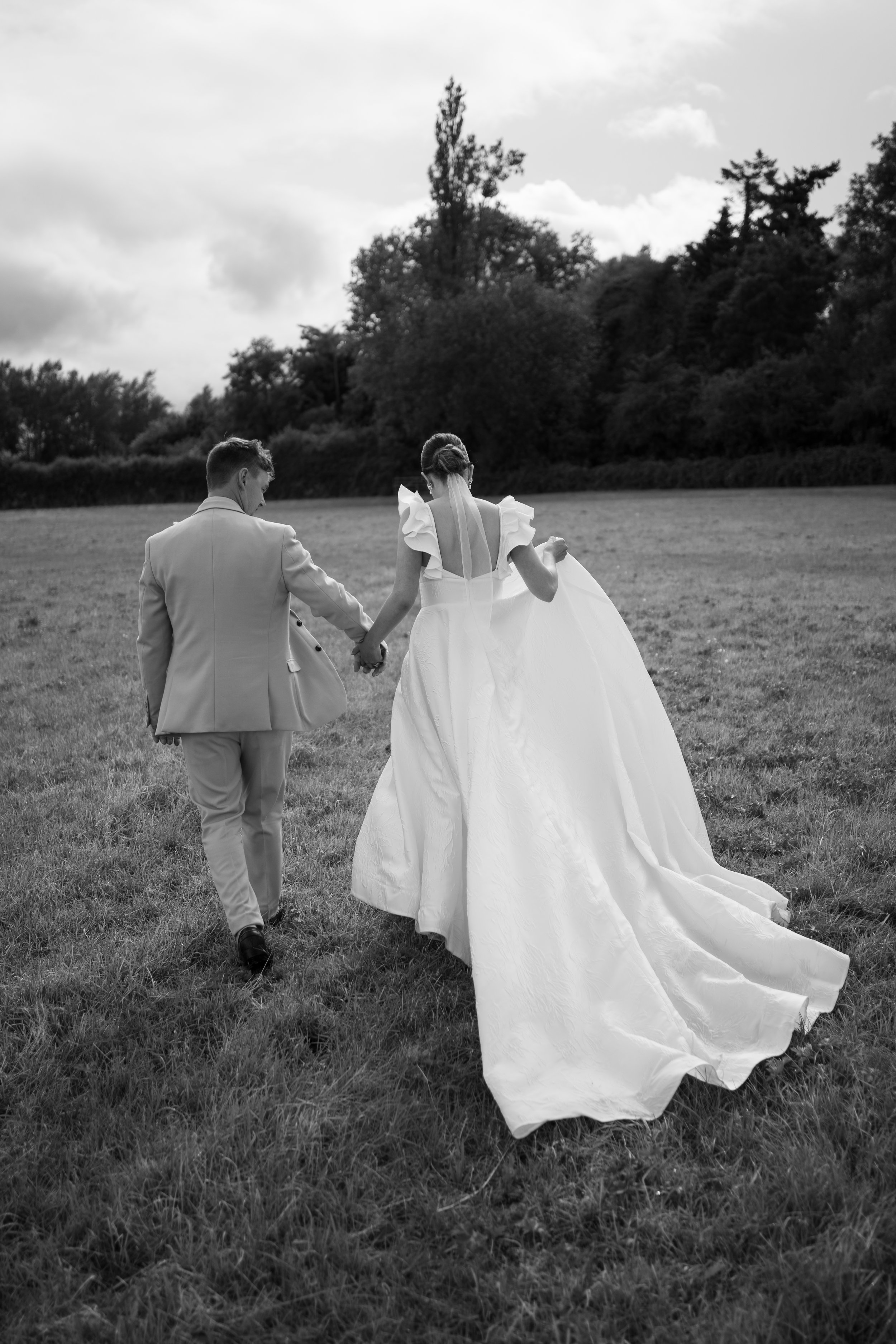 Bride and groom walking hand in hand across a grassy field, with the bride in a flowing wedding dress and veil, and the groom in a suit.