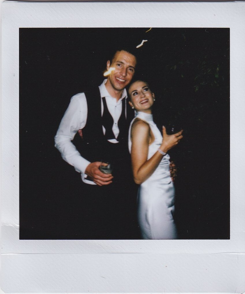 A couple dressed in formal attire posing together, smiling, and holding drinks against a dark background.