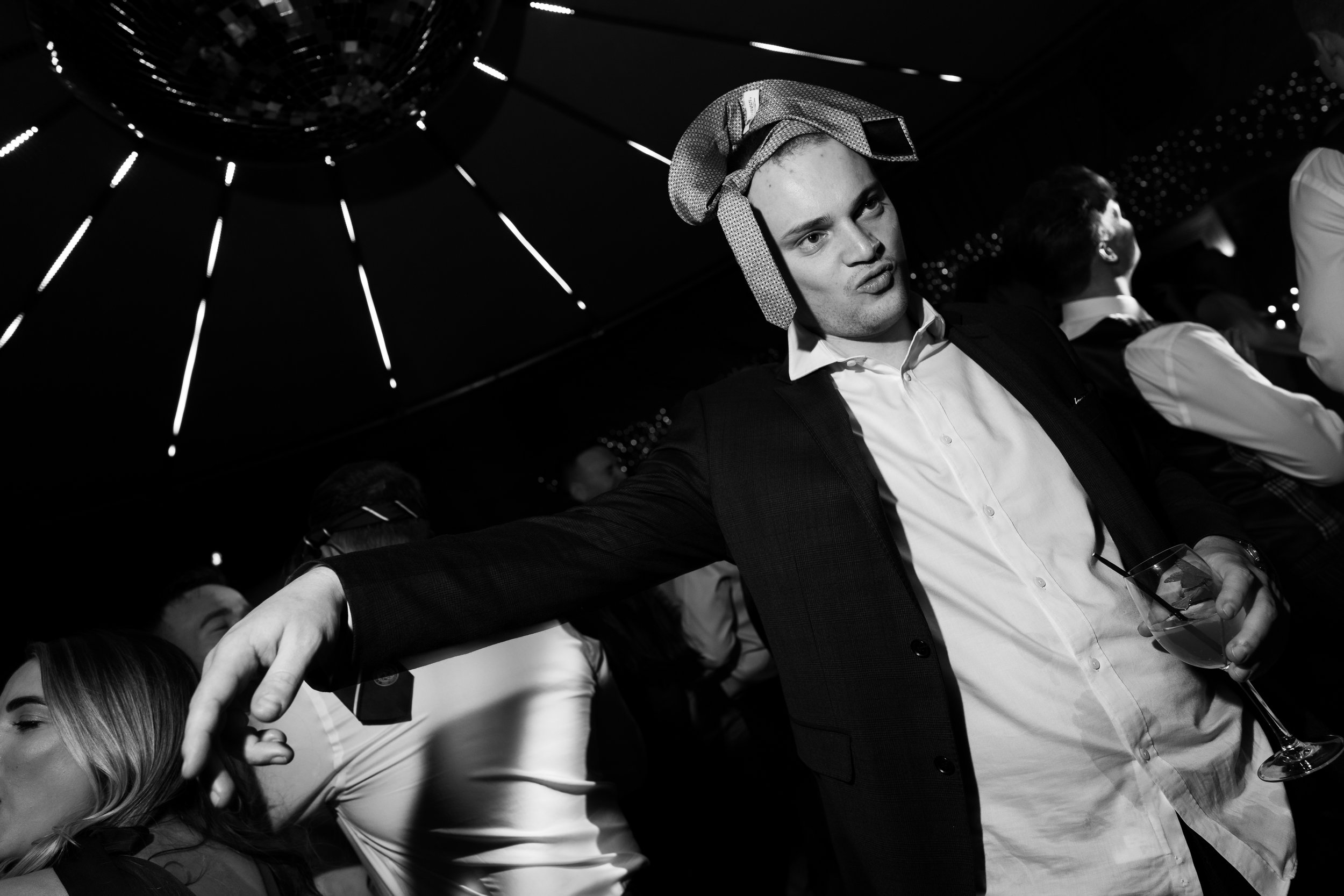 Black and white photo of a man in a suit, with a tie on his head, holding a drink, at a party.