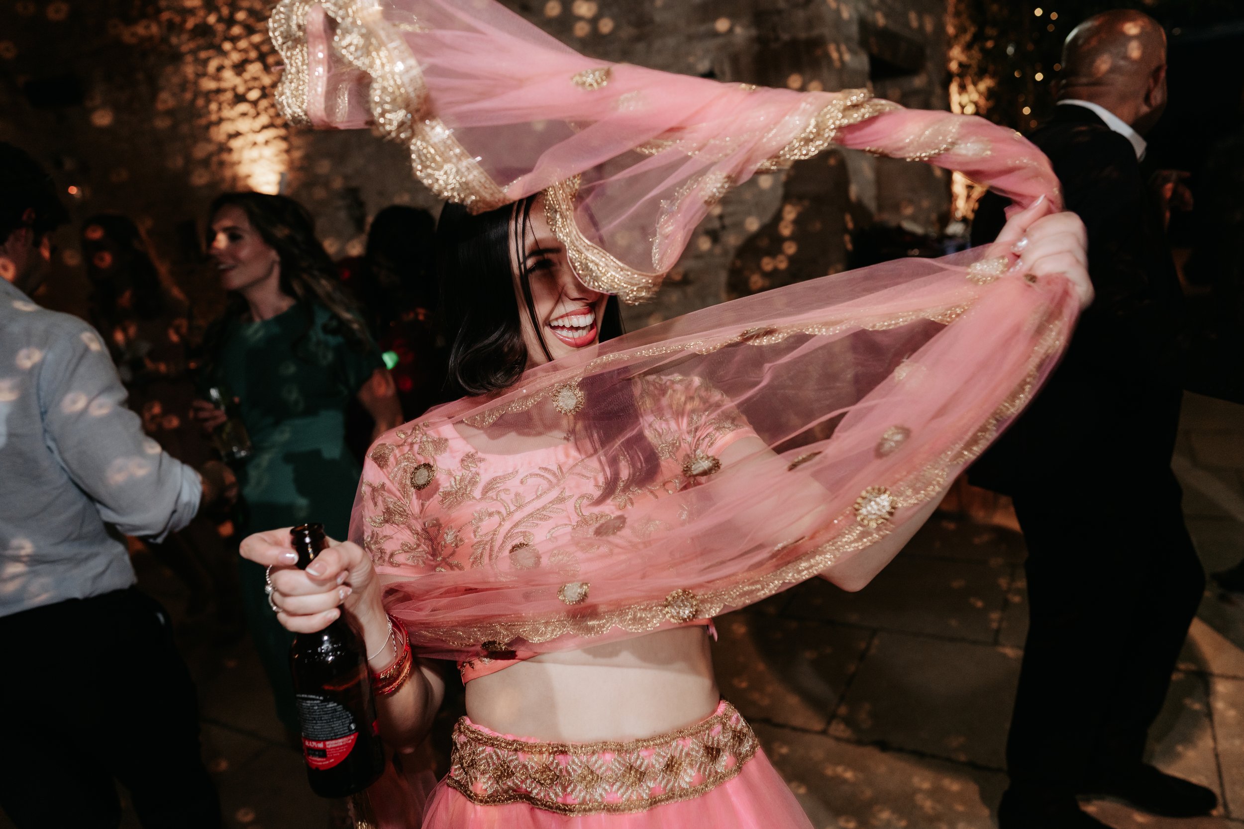 Woman dancing with pink scarf at a party, holding a beer bottle.