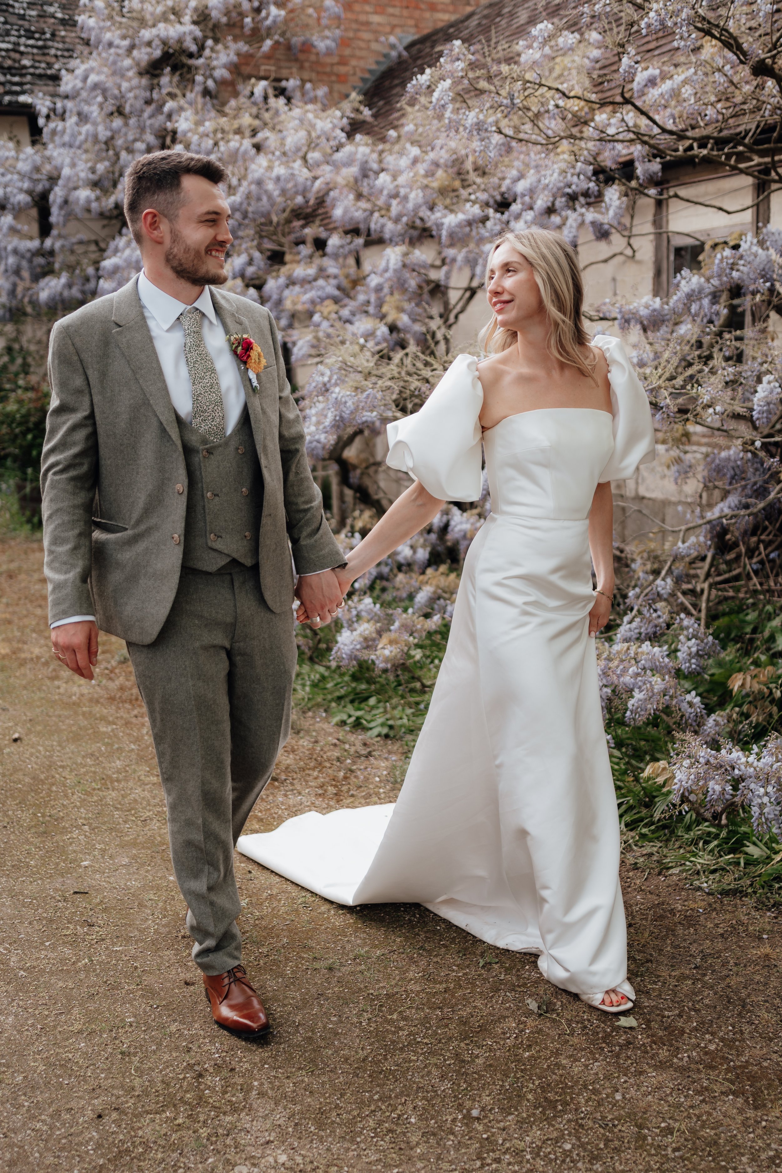 A couple in wedding attire, holding hands and smiling at each other, surrounded by flowering wisteria.