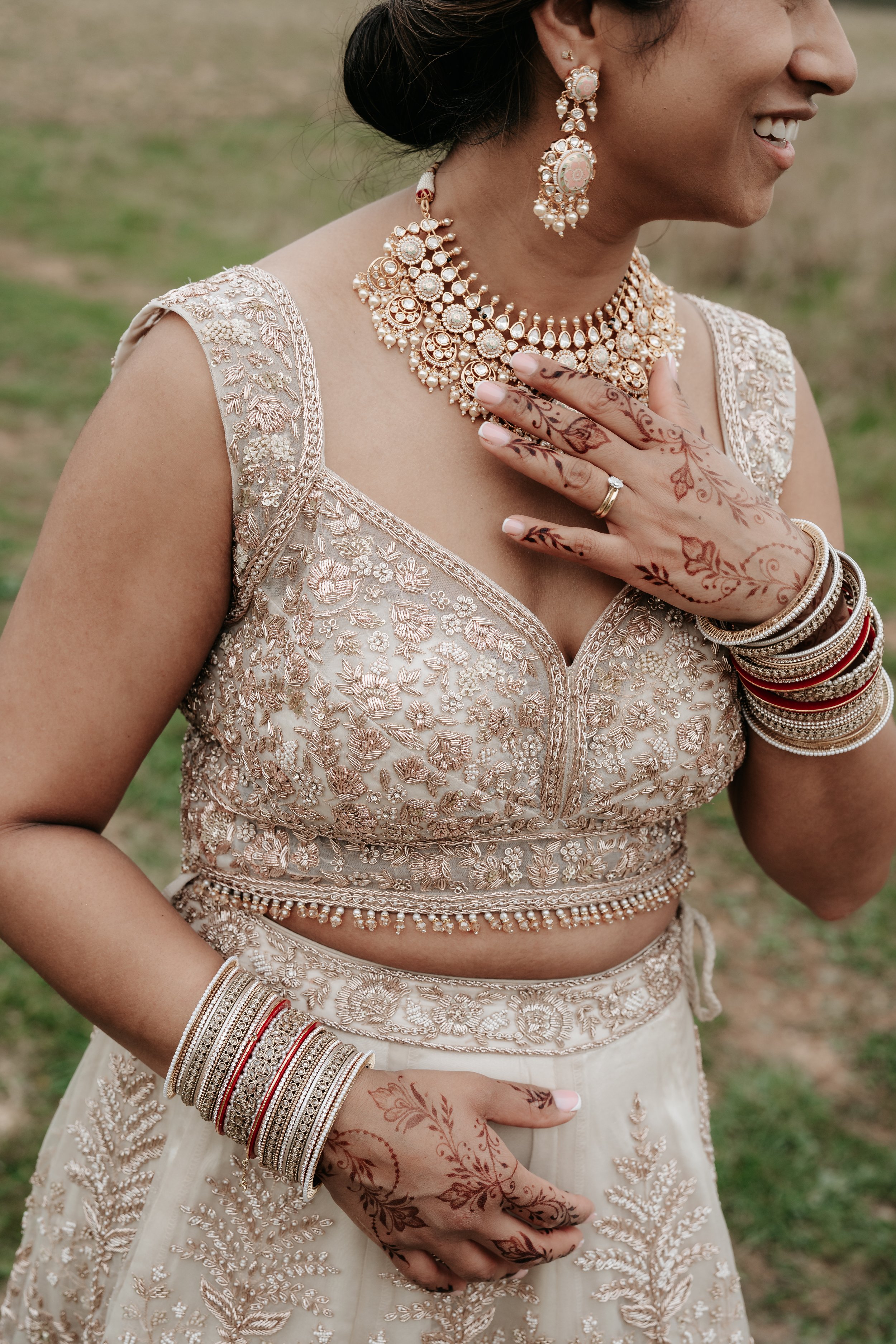 A woman wearing an intricately embroidered bridal outfit with gold jewelry, including a necklace and earrings, and a gold ring. Her hands feature henna designs and she is wearing several bangles. The background shows a grassy area.