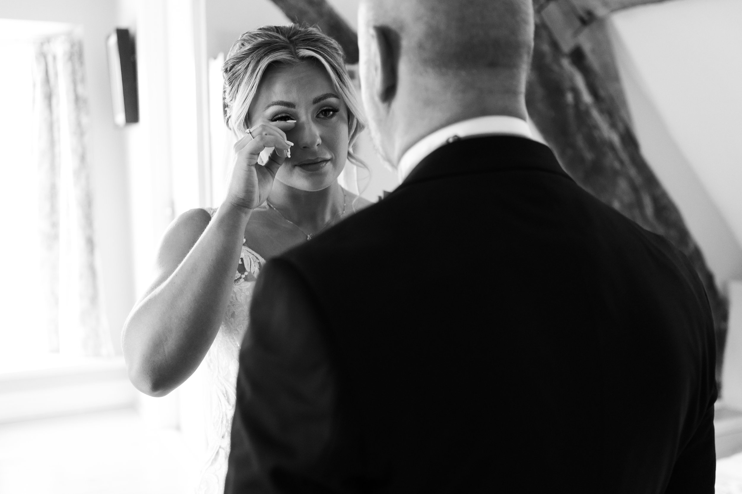 Black and white image of a bride wiping tears while facing a man in a suit, indoors.