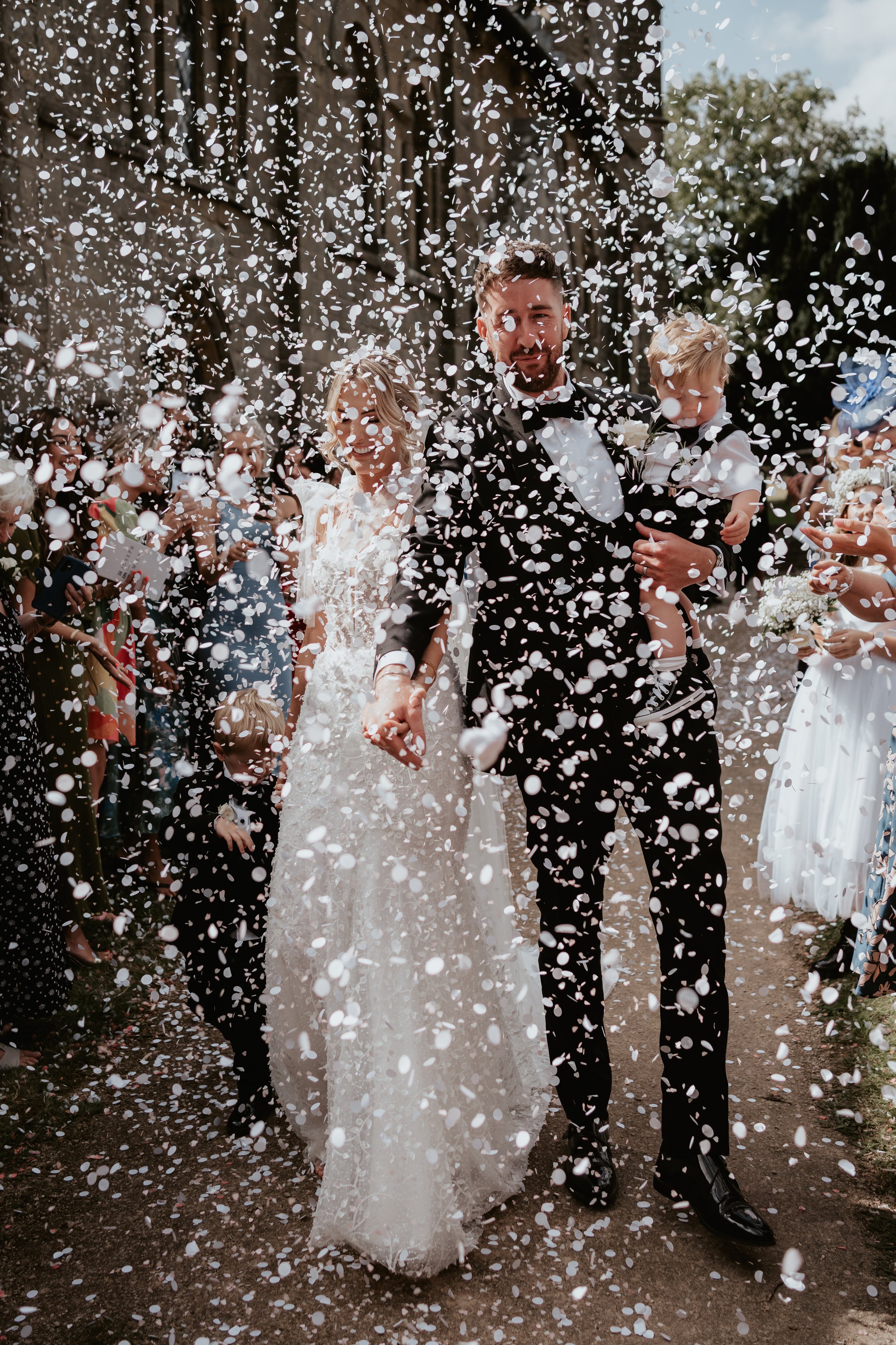 Bride and groom walking with children in a shower of confetti outside a building during a wedding celebration.