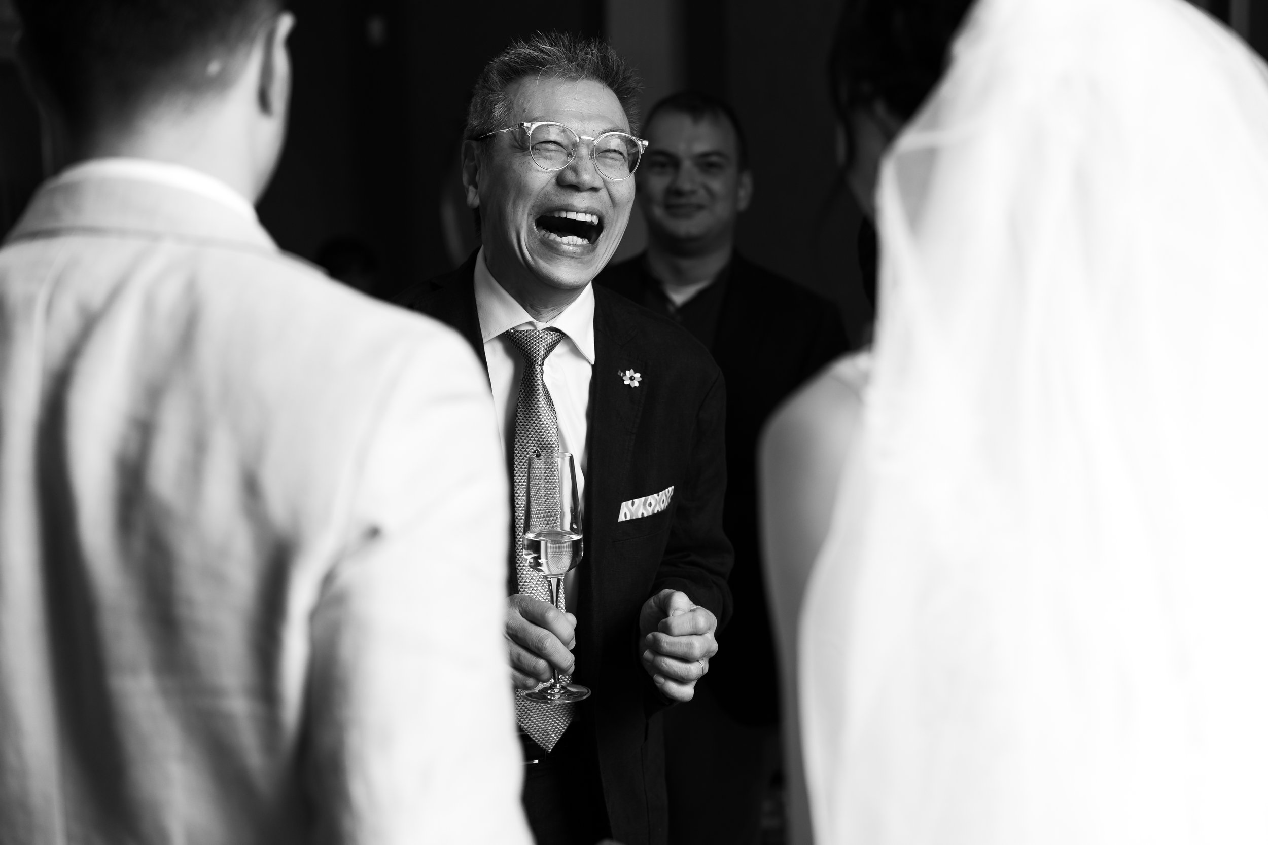 A black and white photo of a man in glasses, laughing while holding a champagne glass, with a bride and groom partially visible in the foreground.