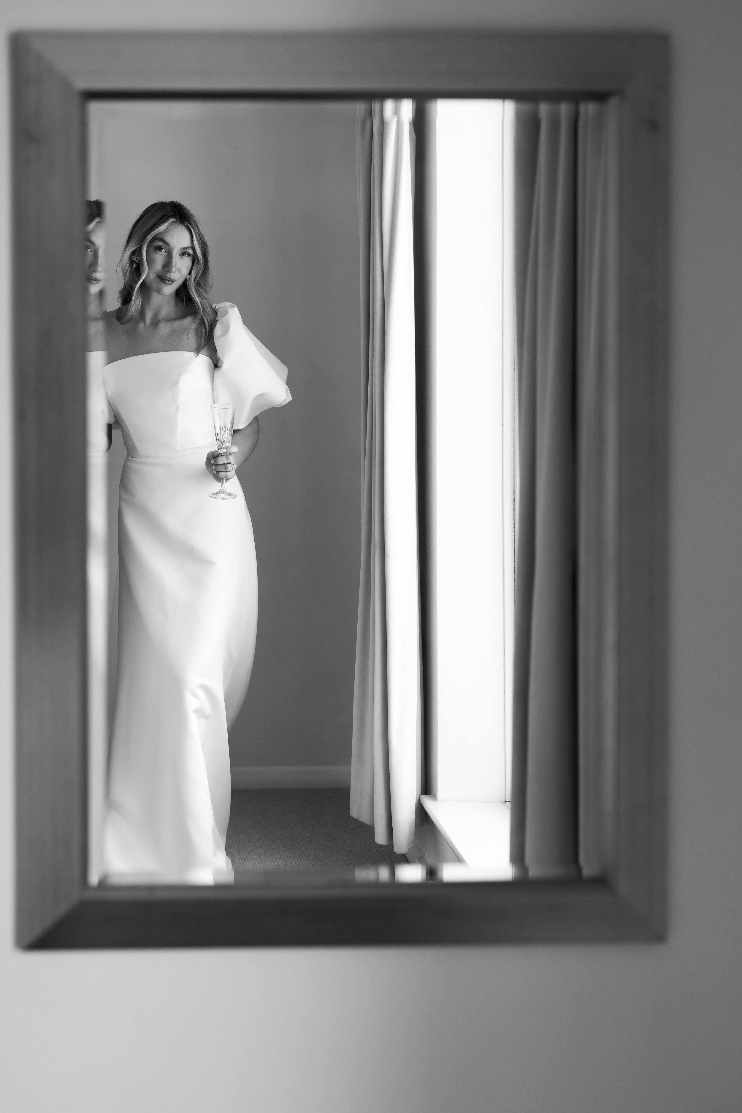 Black and white reflection of a bride in a mirror, wearing a white gown and holding a champagne glass.