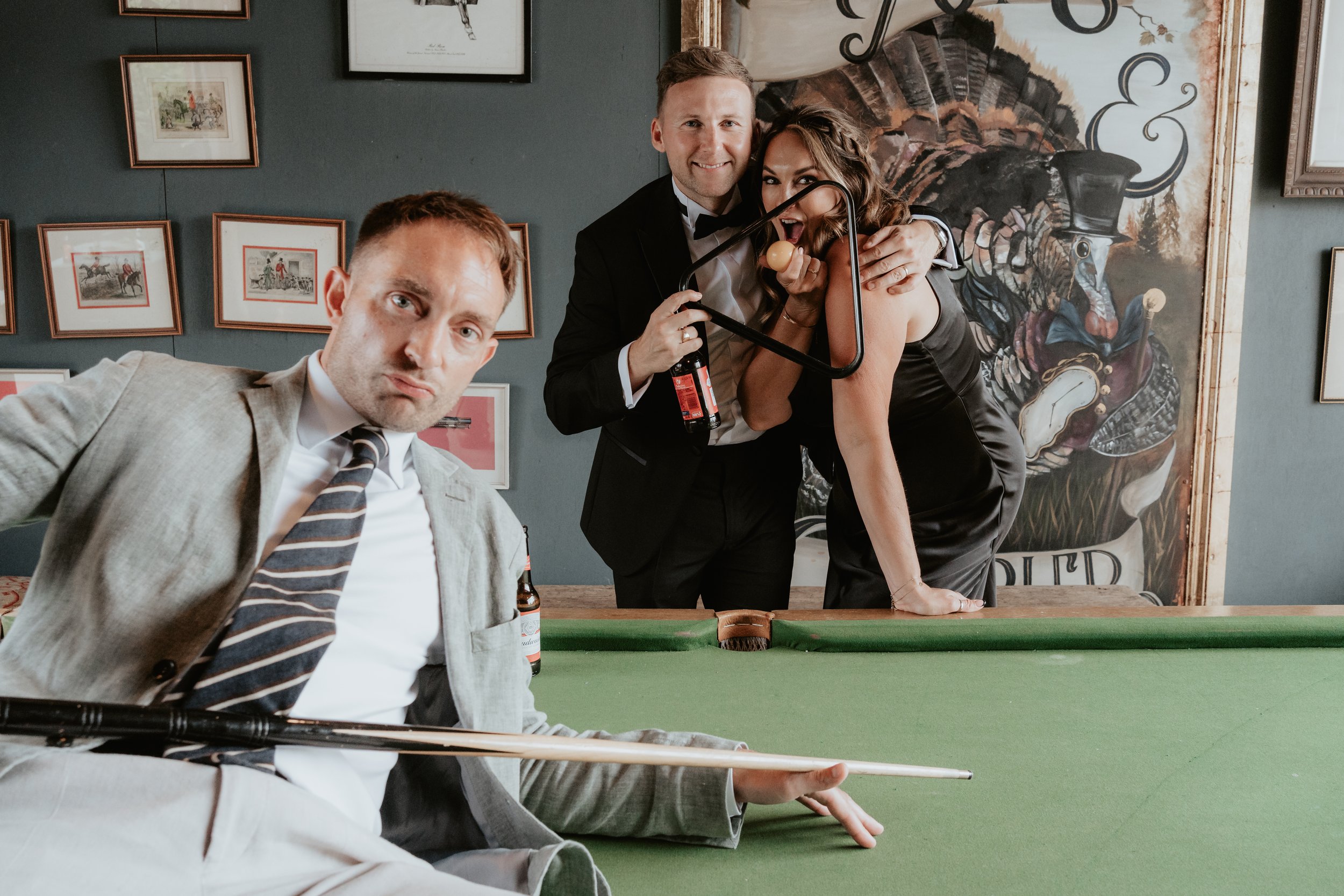 Three people posing playfully around a pool table, one in a gray suit leaning with a cue, two in formal wear smiling, one holding a pool triangle.