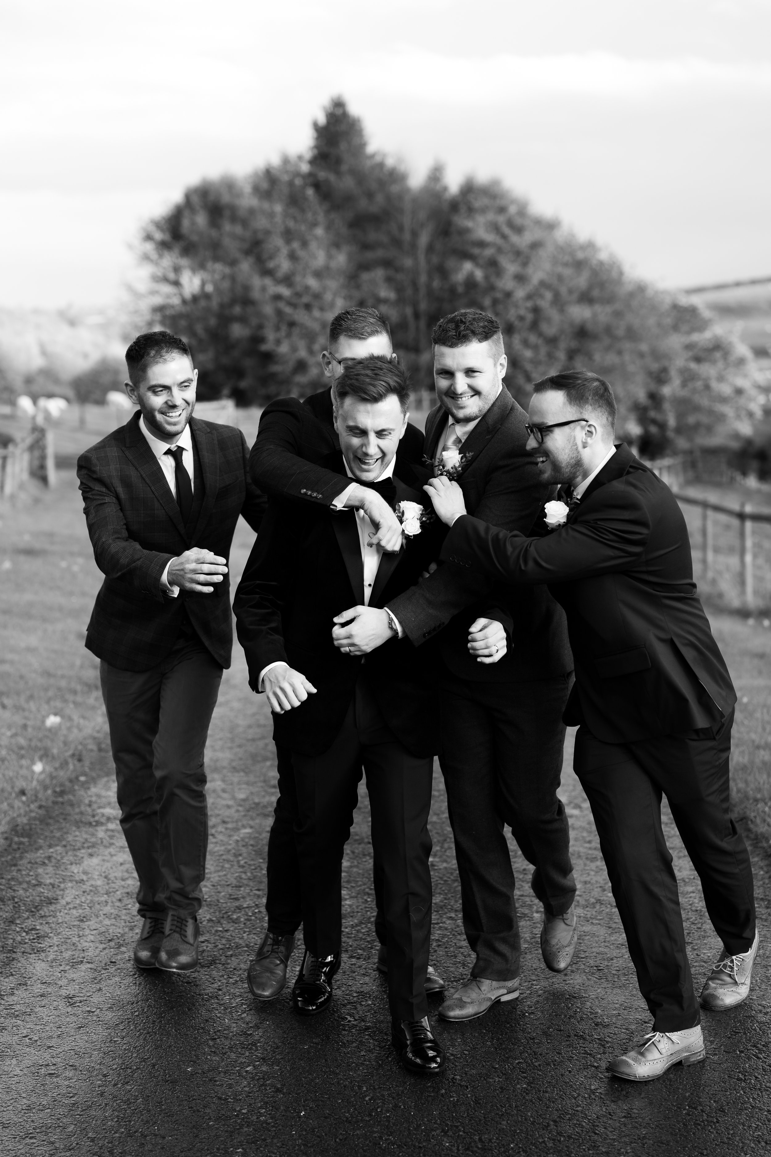 Group of groomsmen in suits laughing and having fun outdoors on a path, with trees in the background.