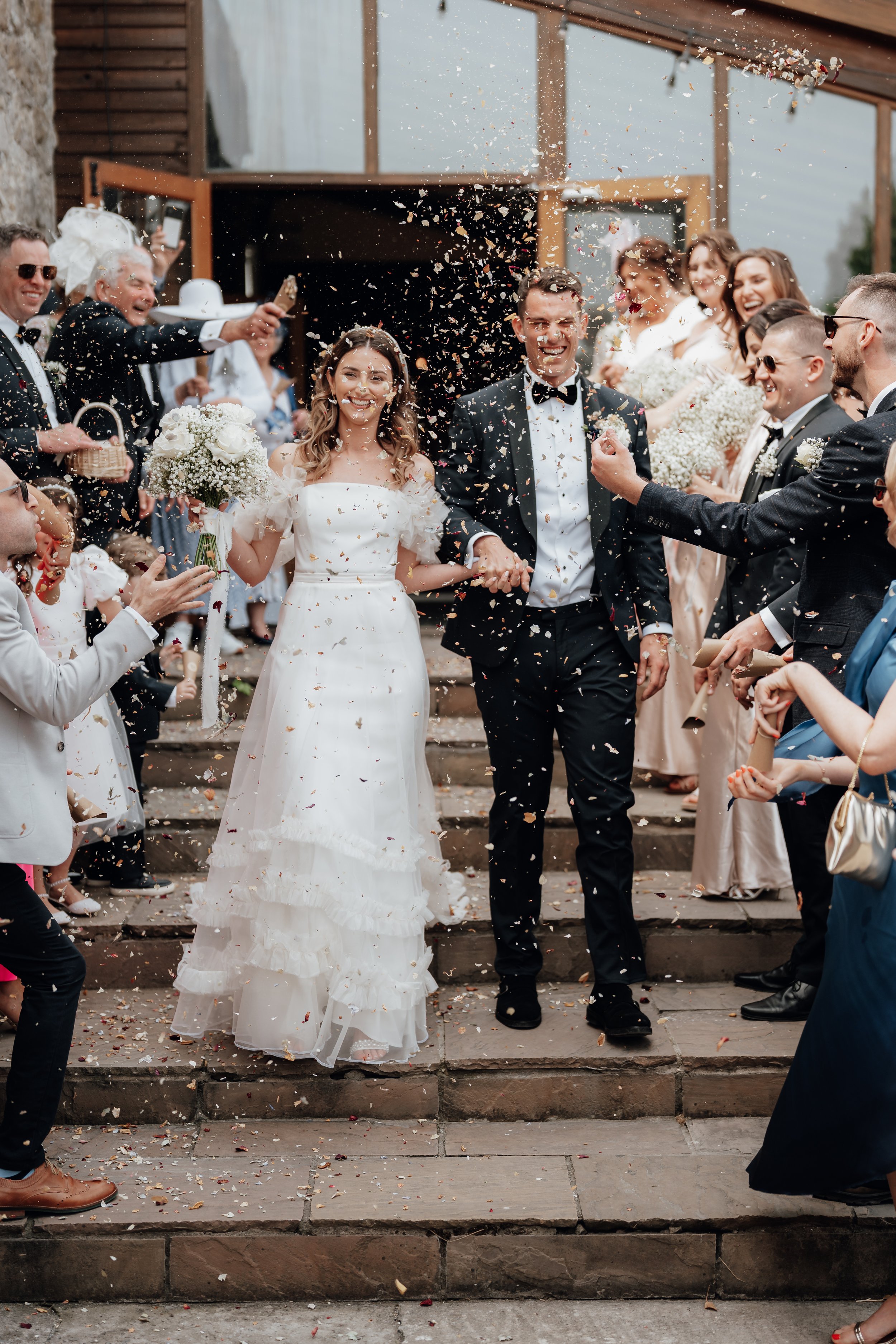 A bride and groom exiting a building, smiling, with confetti being thrown over them by a crowd of well-dressed guests on a staircase.