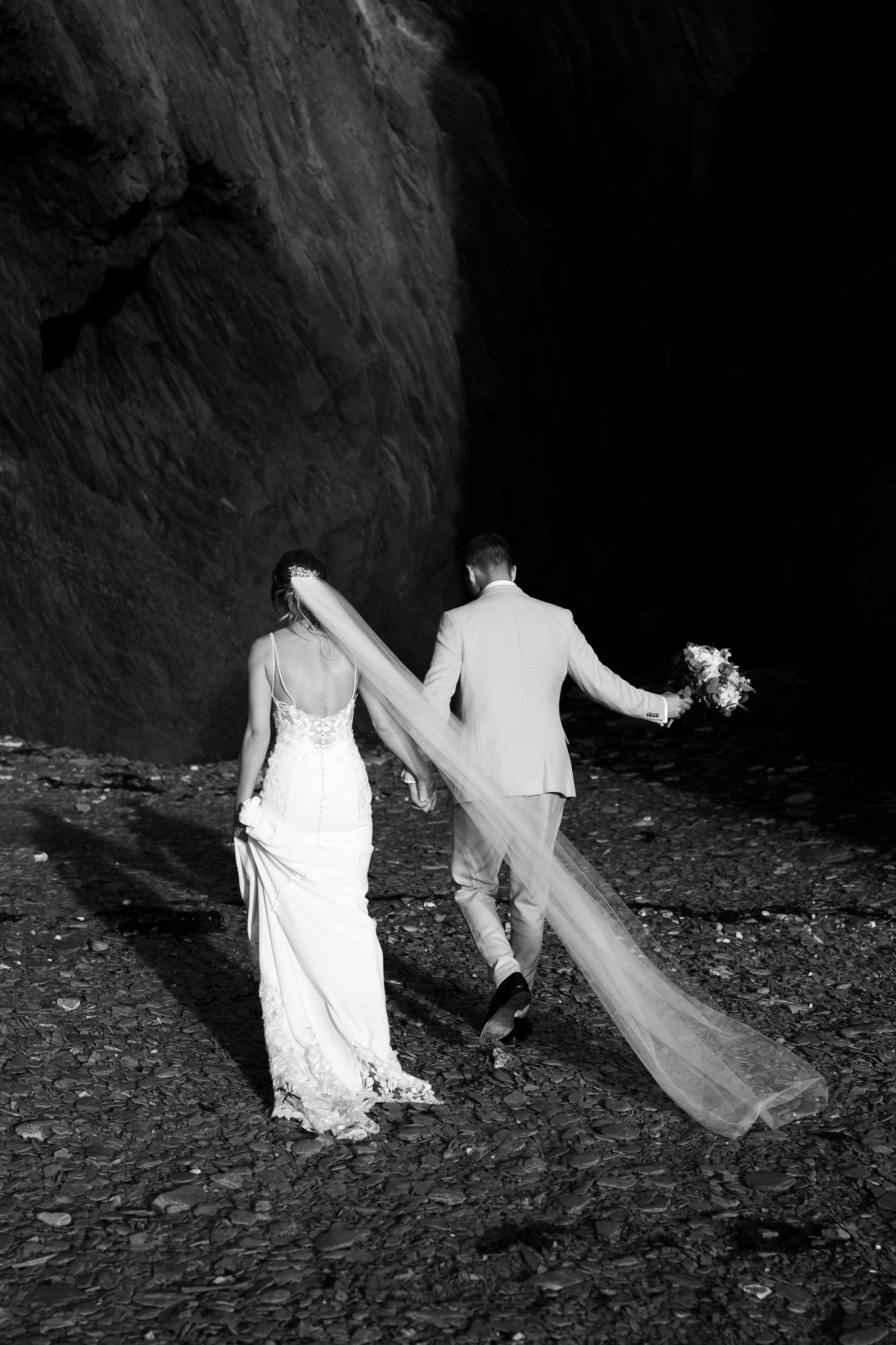 Bride and groom walking on a rocky path in a cave, bride wearing a long veil and holding groom's hand, groom carrying a bouquet. Black and white photo.
