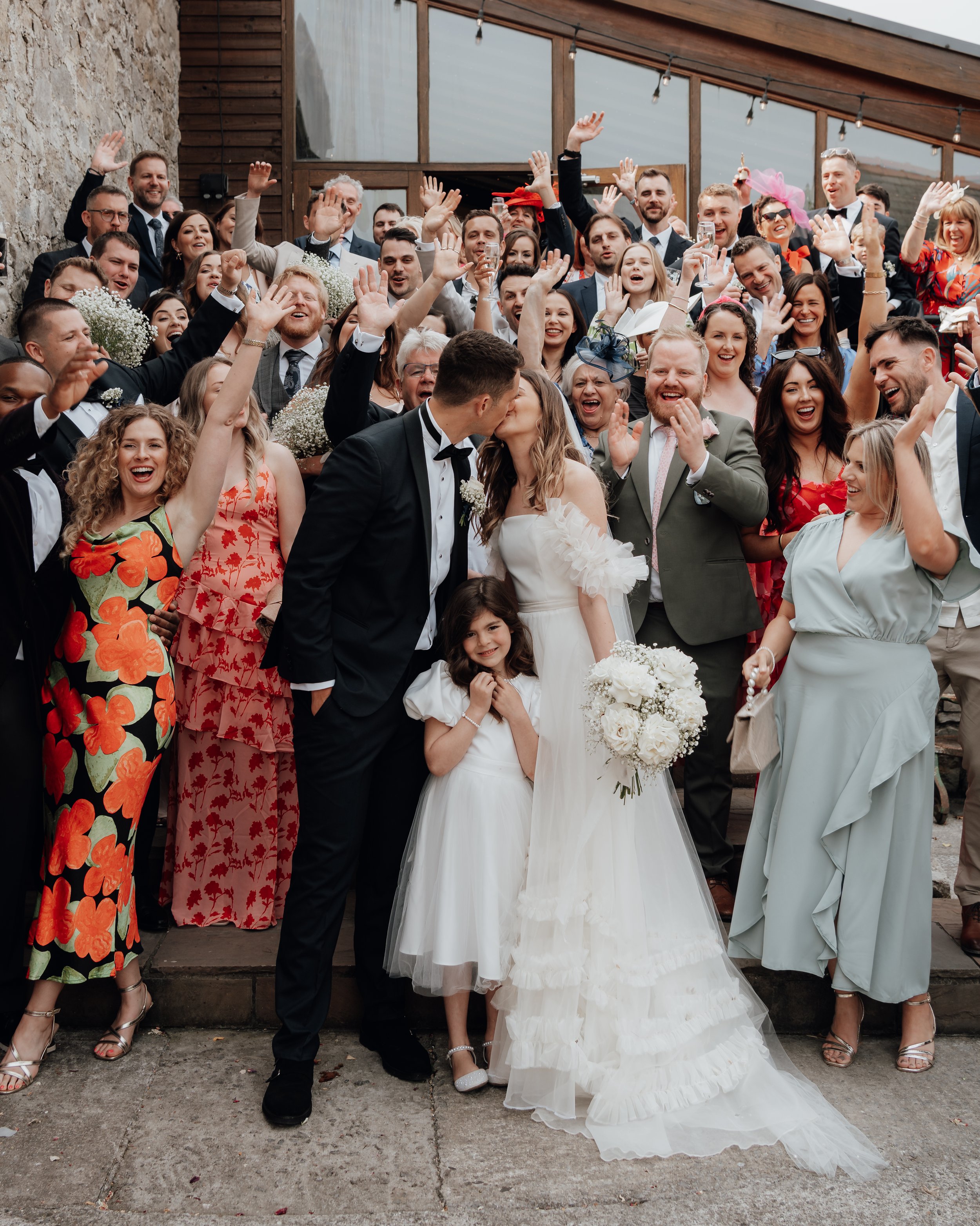 A wedding group photo with a bride and groom kissing, surrounded by guests, including a young girl in a white dress. The guests are smiling, cheering, and waving.