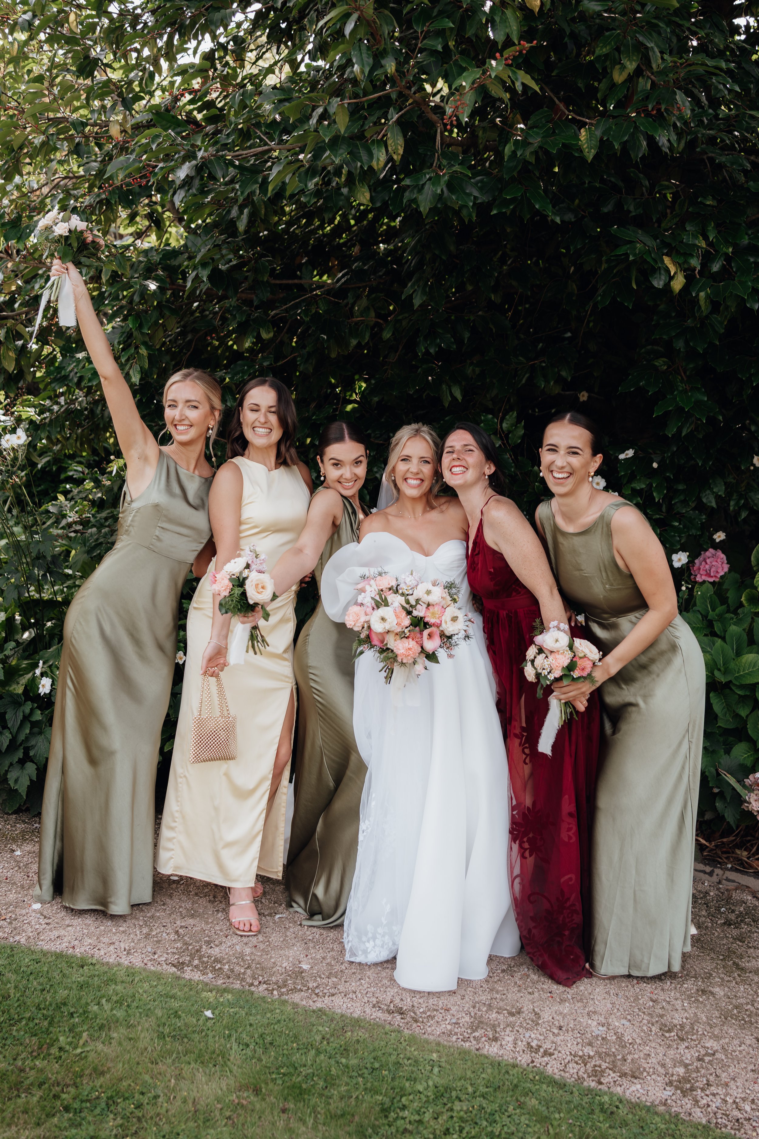 A bride in a white gown and five bridesmaids wearing different colored dresses pose together outdoors, each holding bouquets of flowers. They are smiling and standing in front of green foliage.