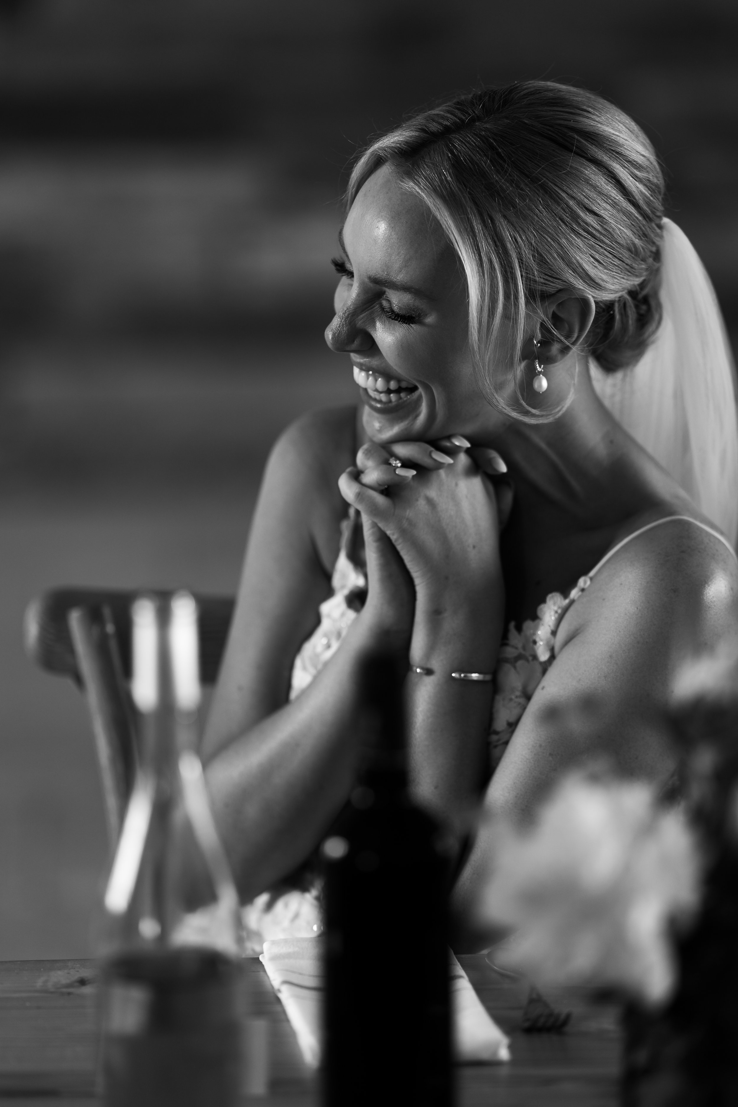 Black and white photo of a smiling woman with her hands clasped, sitting at a table with blurred bottles and flowers in the foreground.