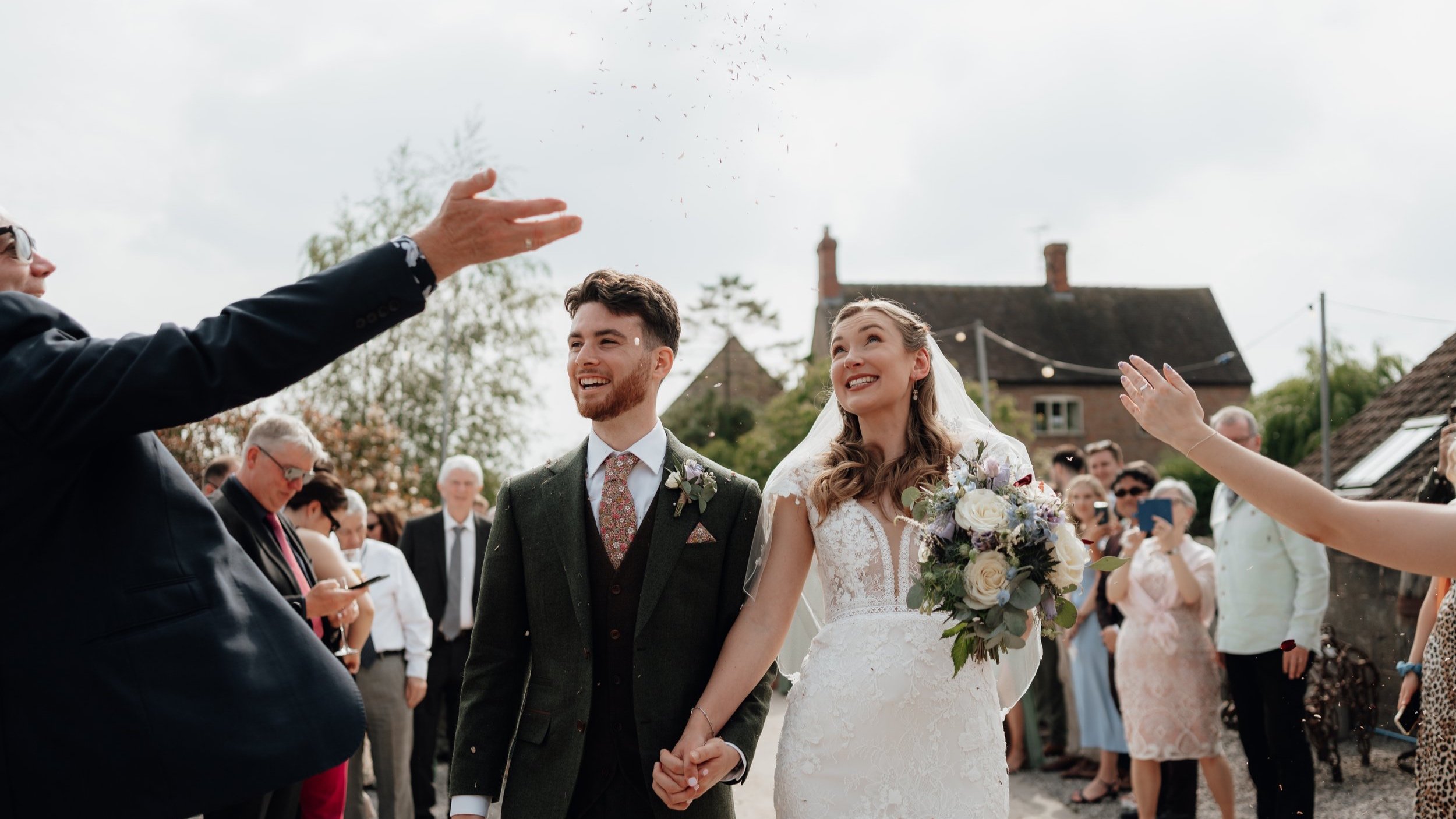 A smiling bride and groom holding hands while guests throw confetti, with a bouquet in the bride's hand and a crowd in the background.