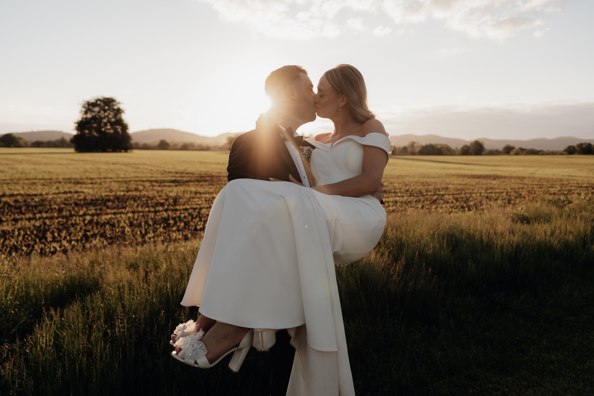 Bride and groom kissing in a field at sunset, with the groom holding the bride in his arms, wearing a white dress and shoes. Background features a tree, grassy field, and mountains.