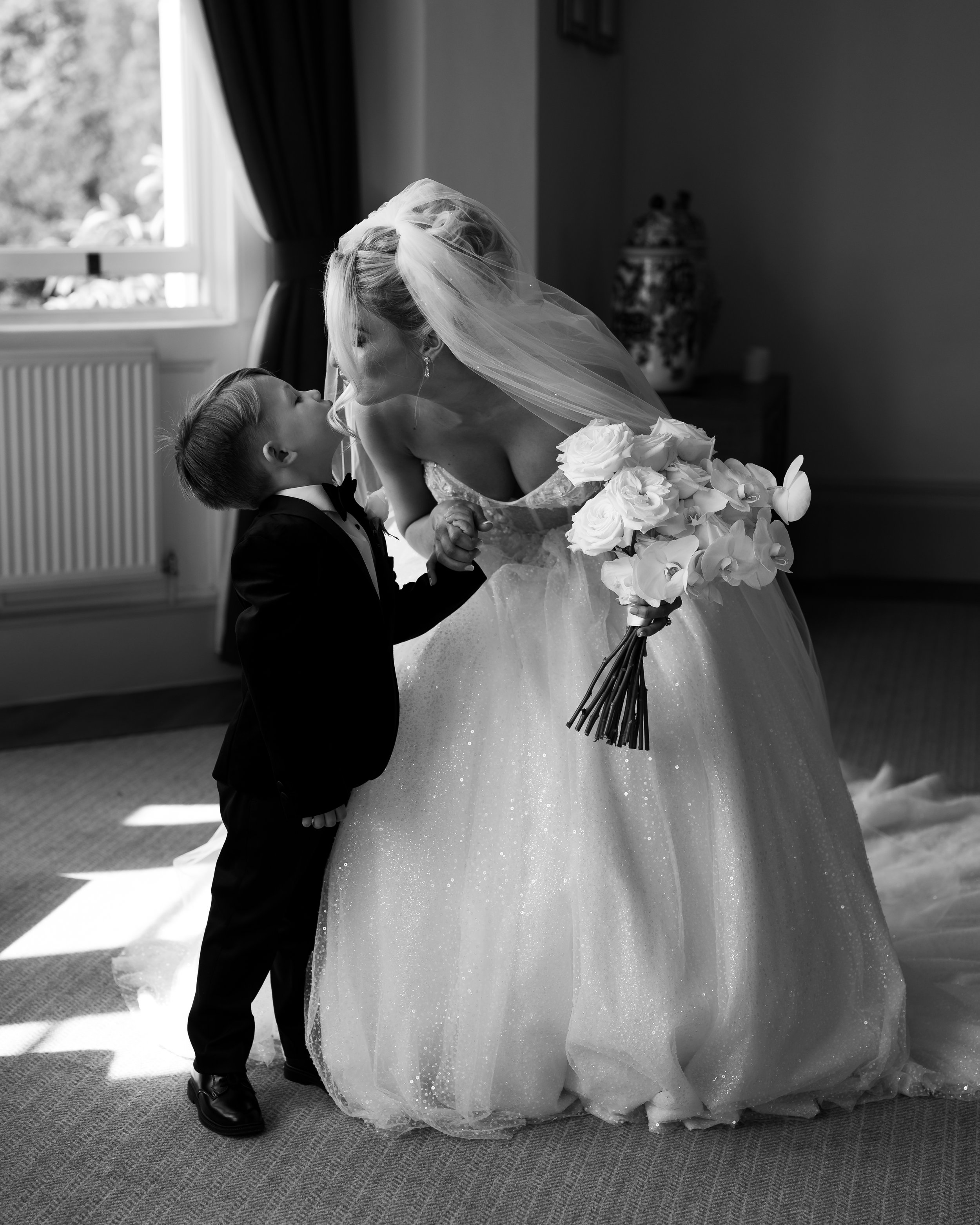 Bride in a wedding dress holding a bouquet, bending down to kiss a young boy in a suit indoors, black and white photo.