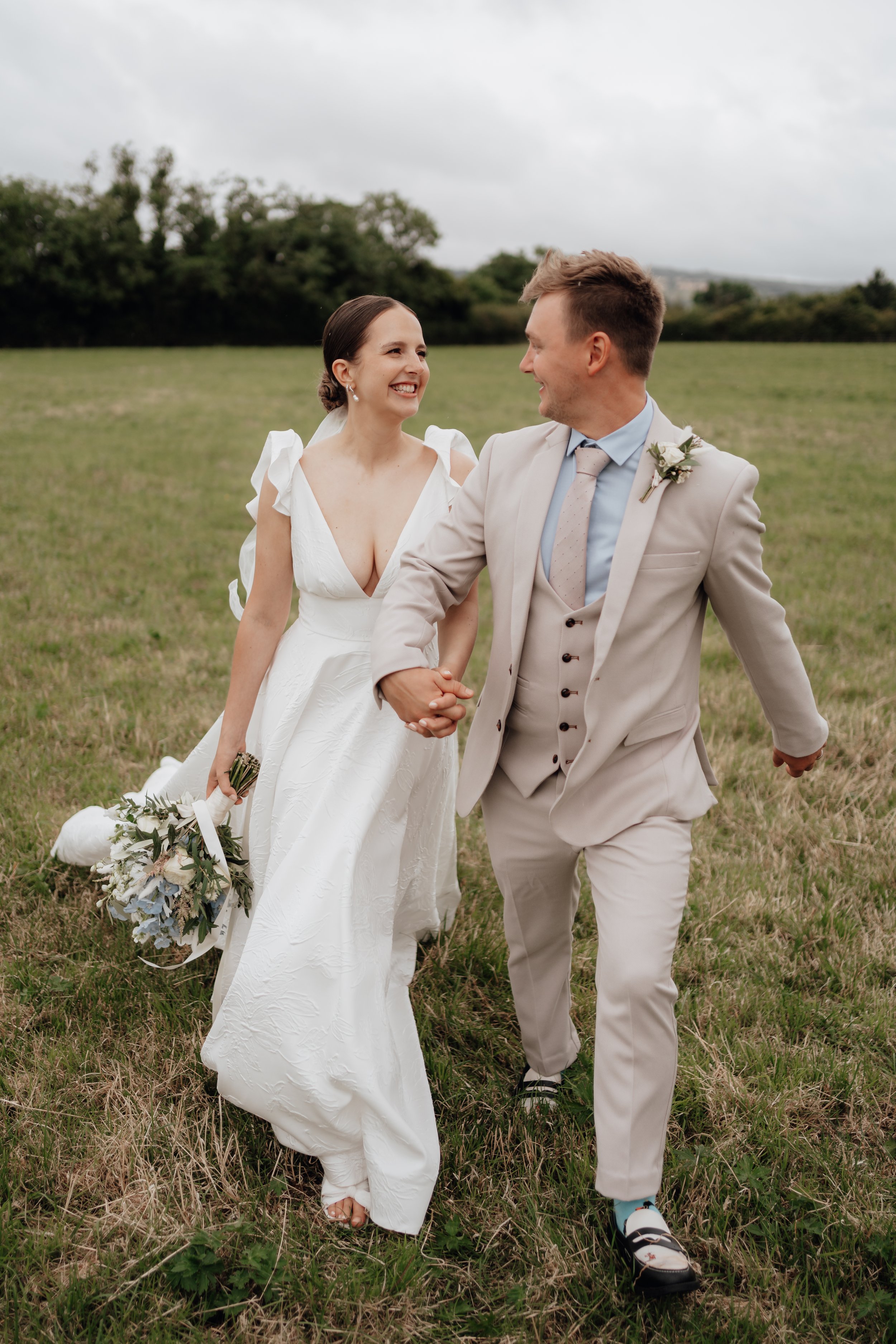 A bride and groom holding hands and smiling in a grassy field, with the bride wearing a white dress and holding a bouquet, and the groom in a light-colored suit.
