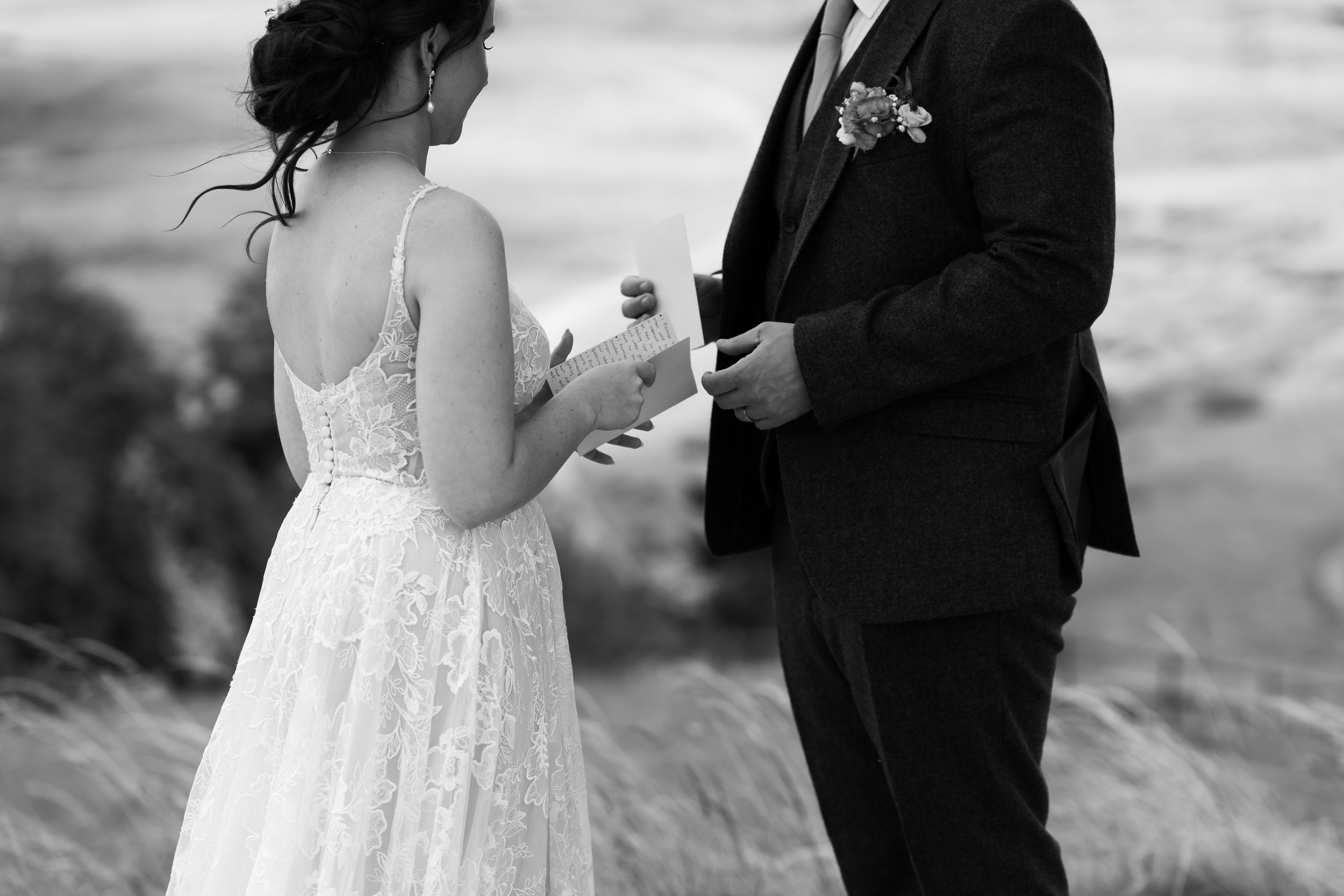 Bride and groom exchanging vows outdoors, woman in lace wedding dress, man in suit with boutonnière, holding notes.