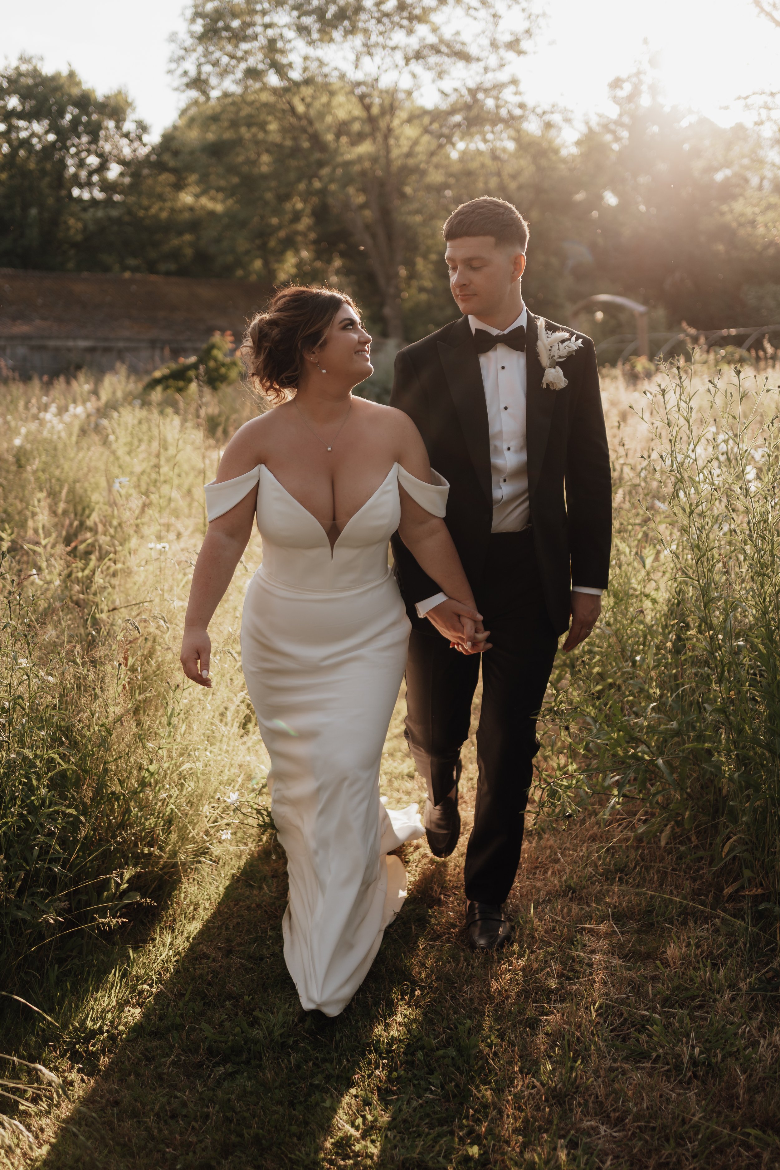 A bride in an off-shoulder wedding dress and a groom in a black tuxedo walk hand in hand through a sunlit field with greenery.