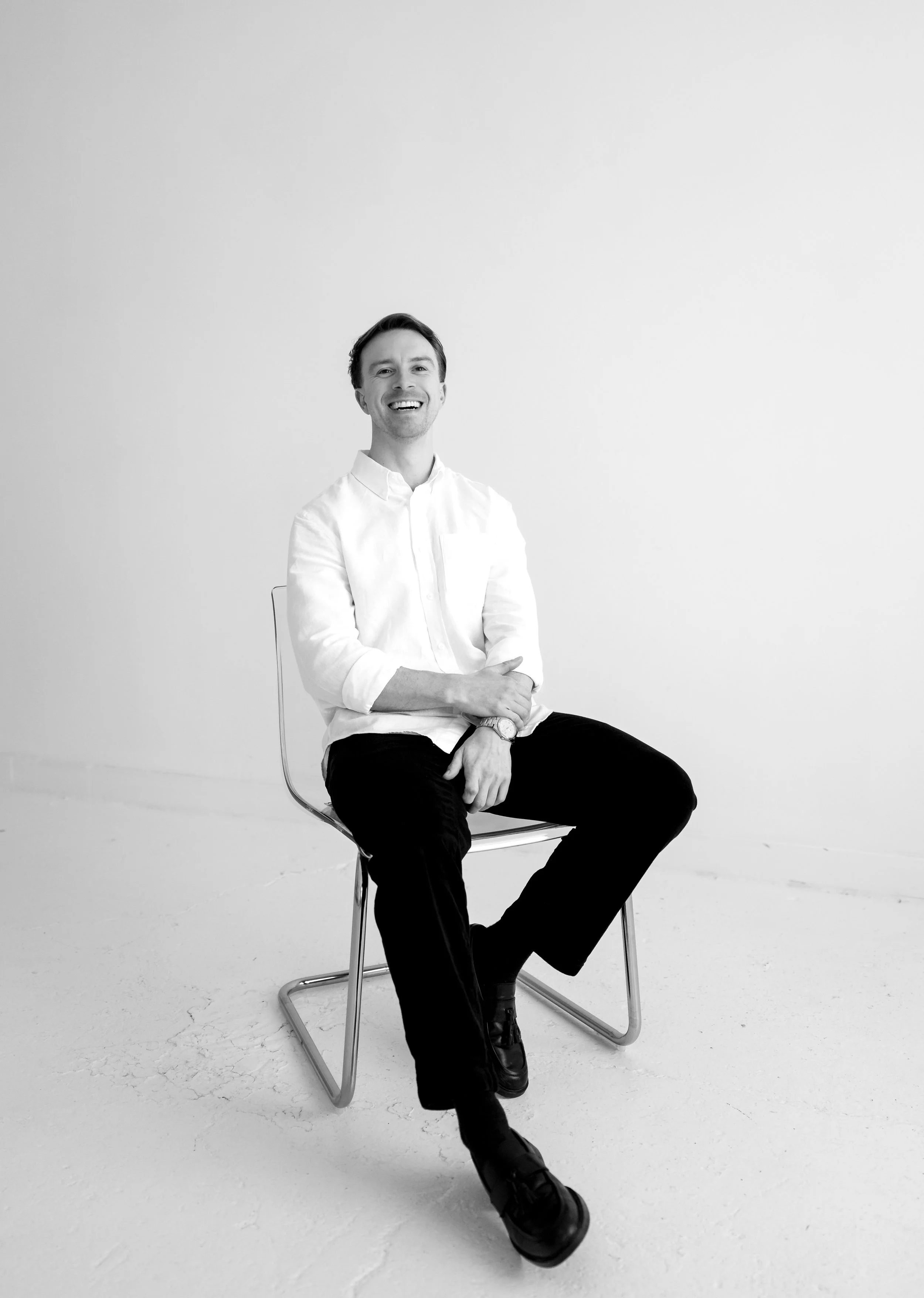 A man with dark hair, wearing a white button-up shirt and black pants, is sitting on a modern wire chair in front of a plain white background. He is smiling and looking at the camera.