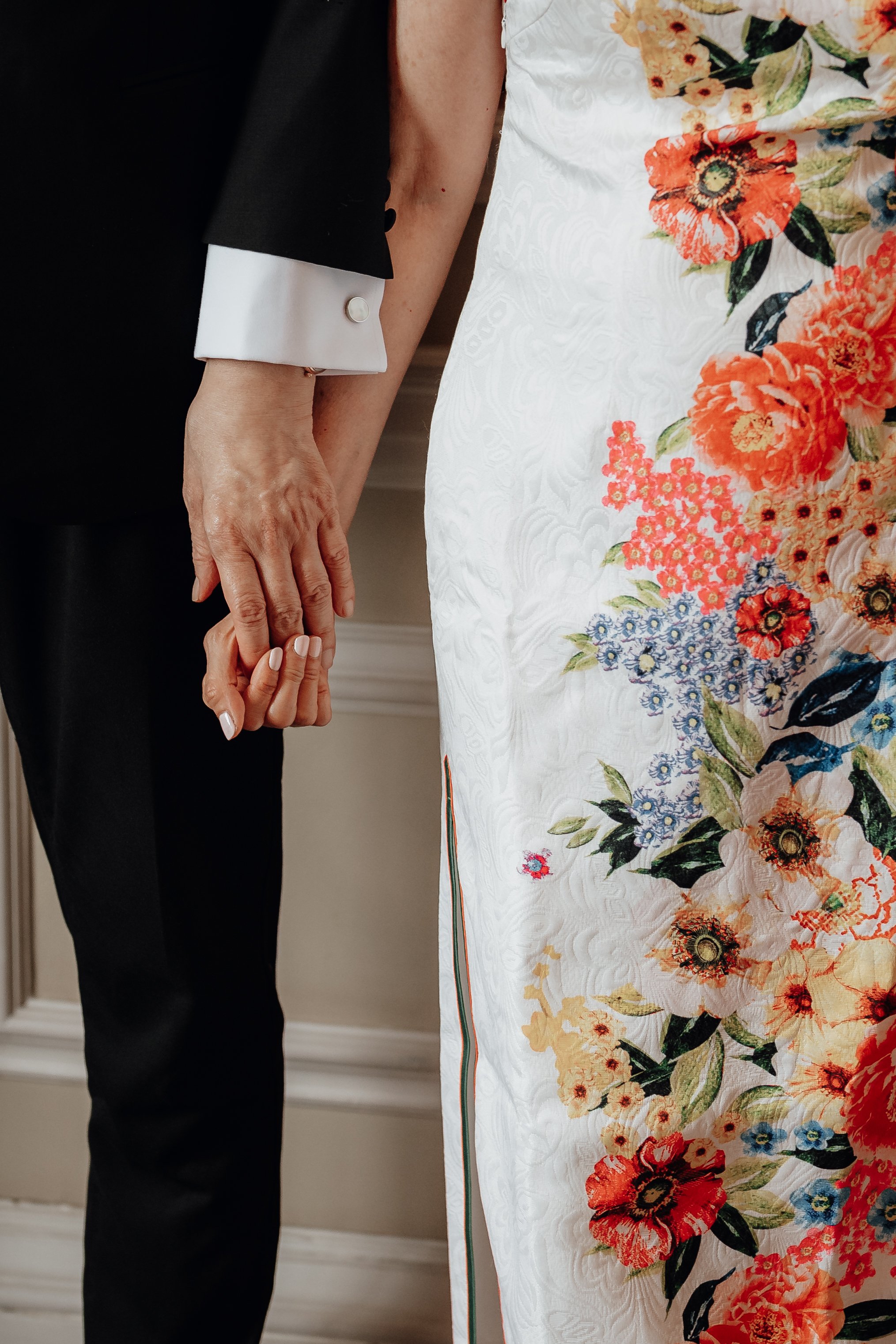 Close-up of a couple holding hands, wearing formal attire; one in a black suit, the other in a white dress with floral embroidery.