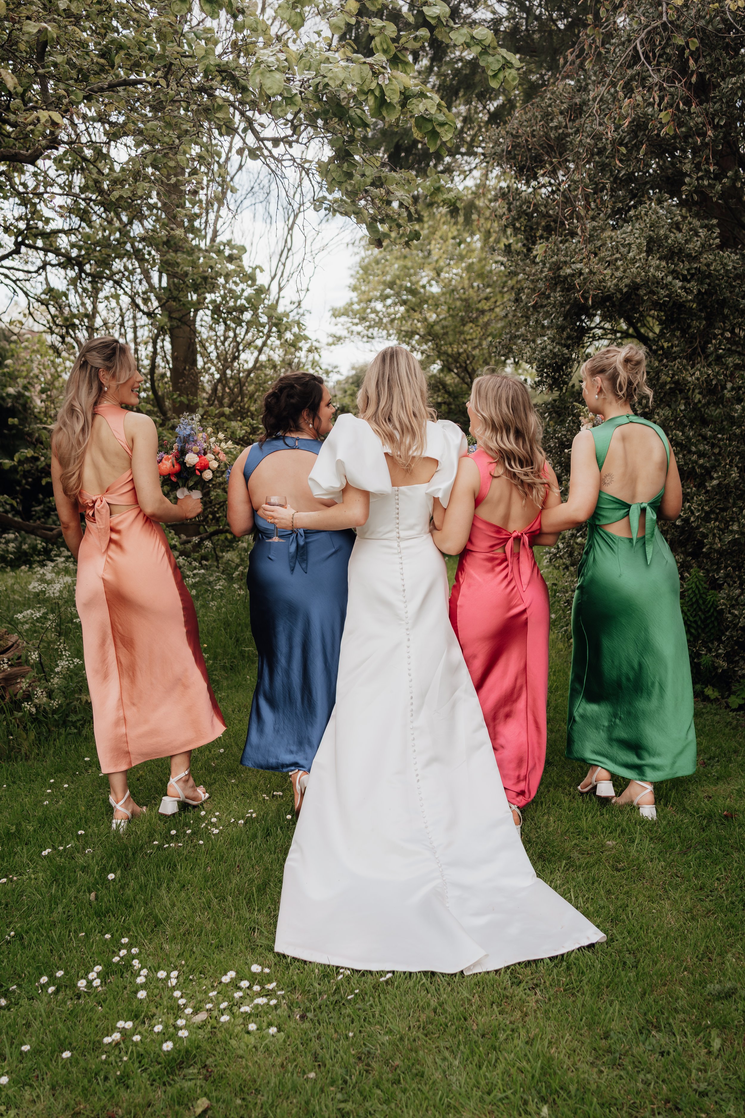 Back view of five women in colorful formal dresses standing on grass in a garden setting.
