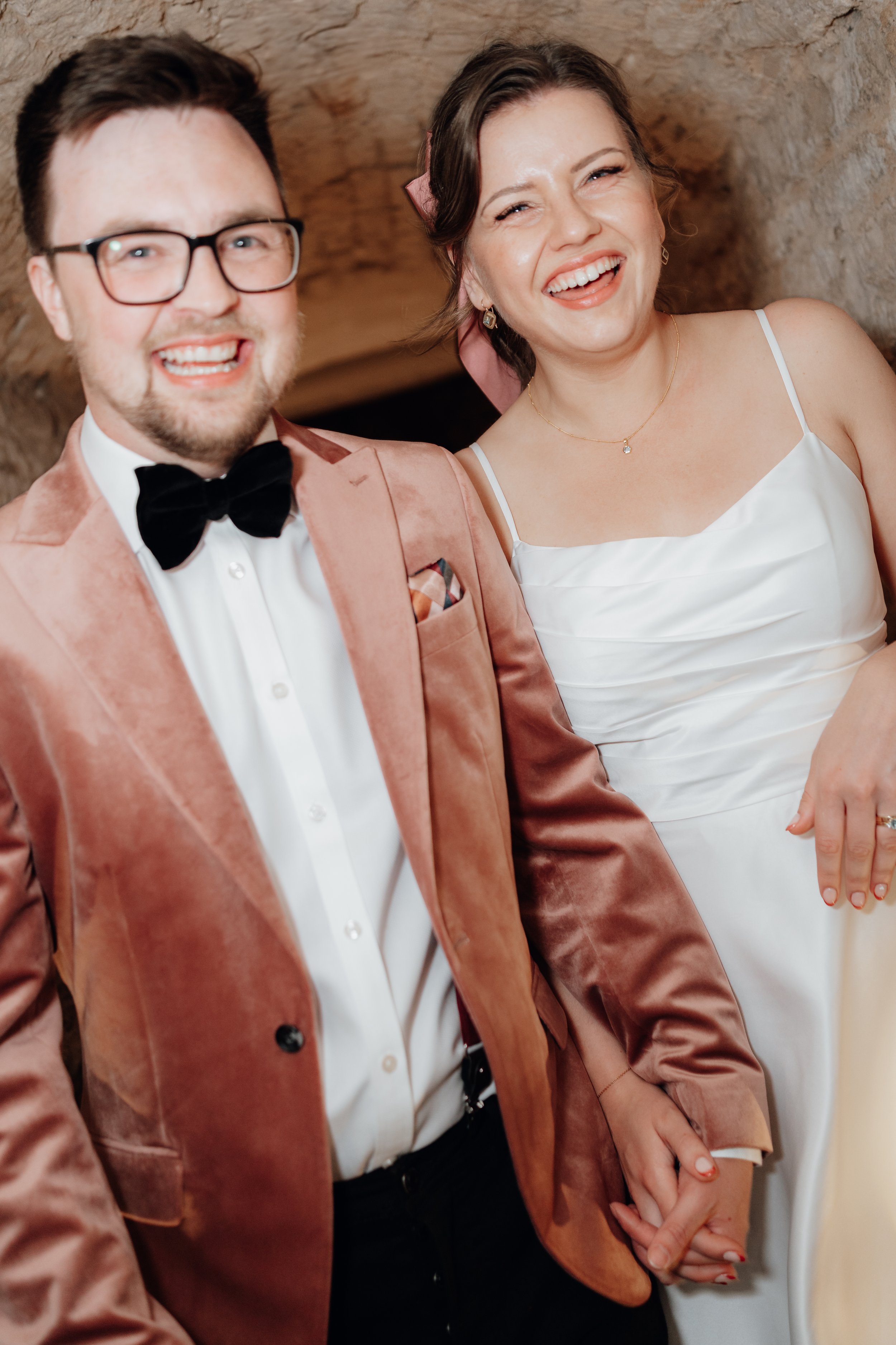 A cheerful couple, with a man in a pink velvet tuxedo and a woman in a white dress, holding hands and smiling.