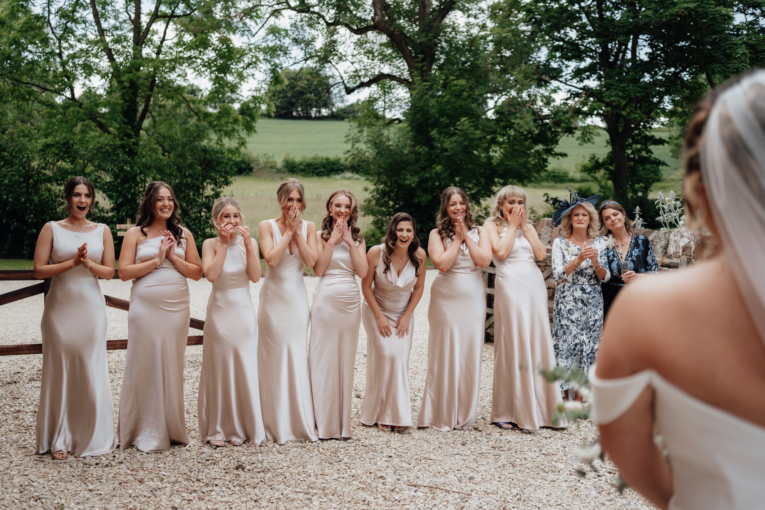 Bridesmaids in long dresses expressing excitement and joy while looking at a bride outside in a garden setting.