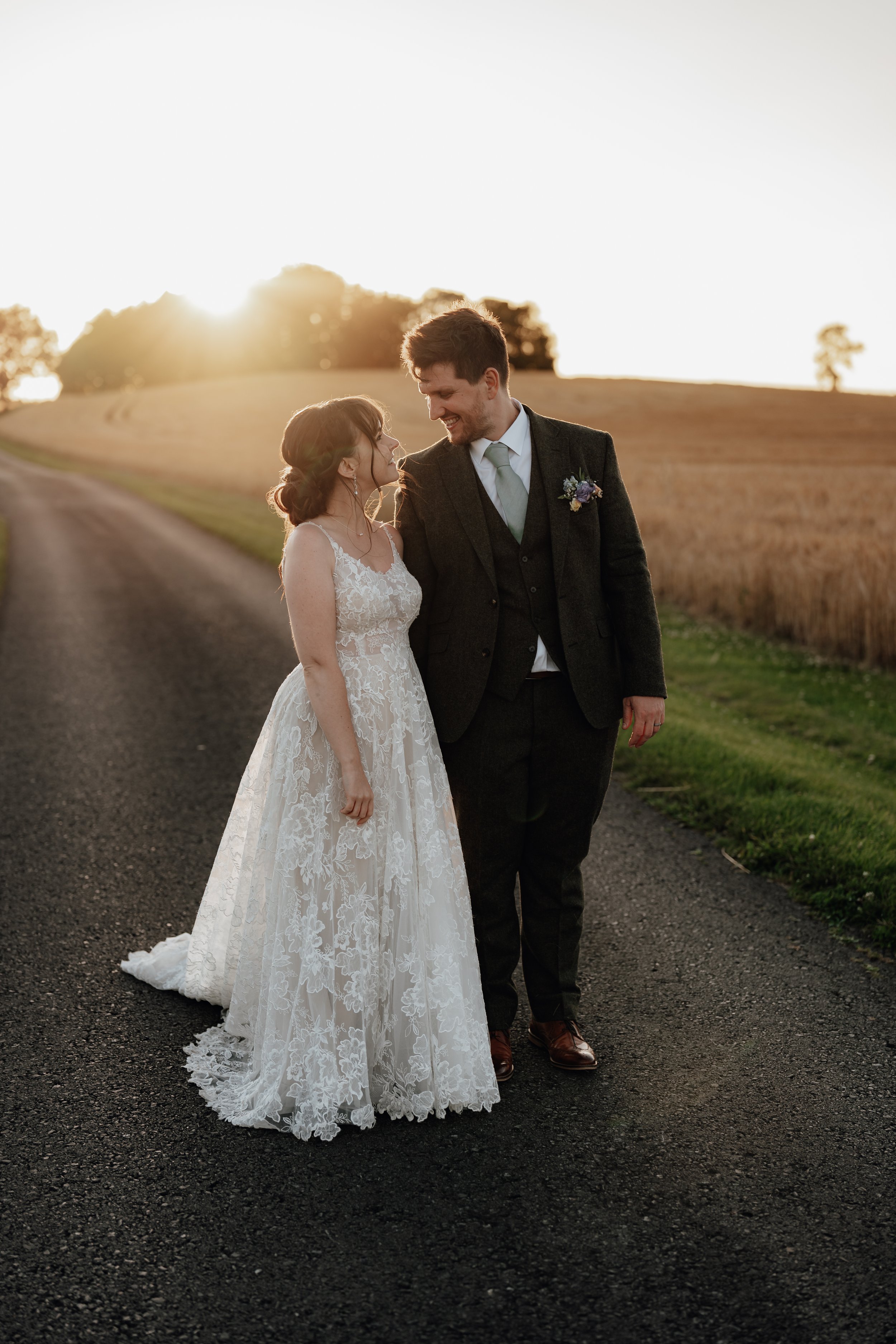 Bride and groom smiling at each other on a country road at sunset, with fields in the background.