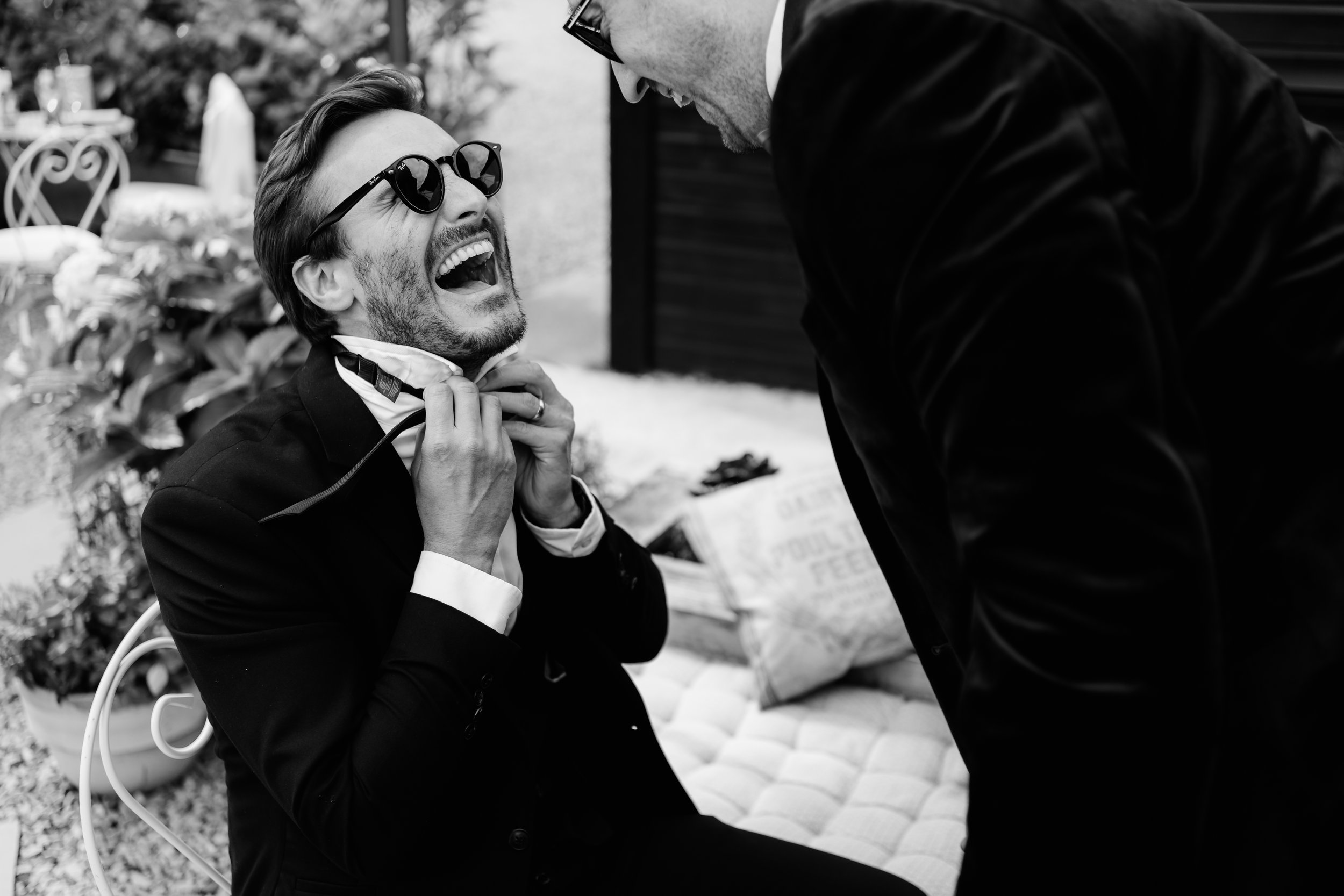 Black and white photo of a man laughing while adjusting tie, wearing sunglasses and a suit, with another person helping, outdoor setting with plants.