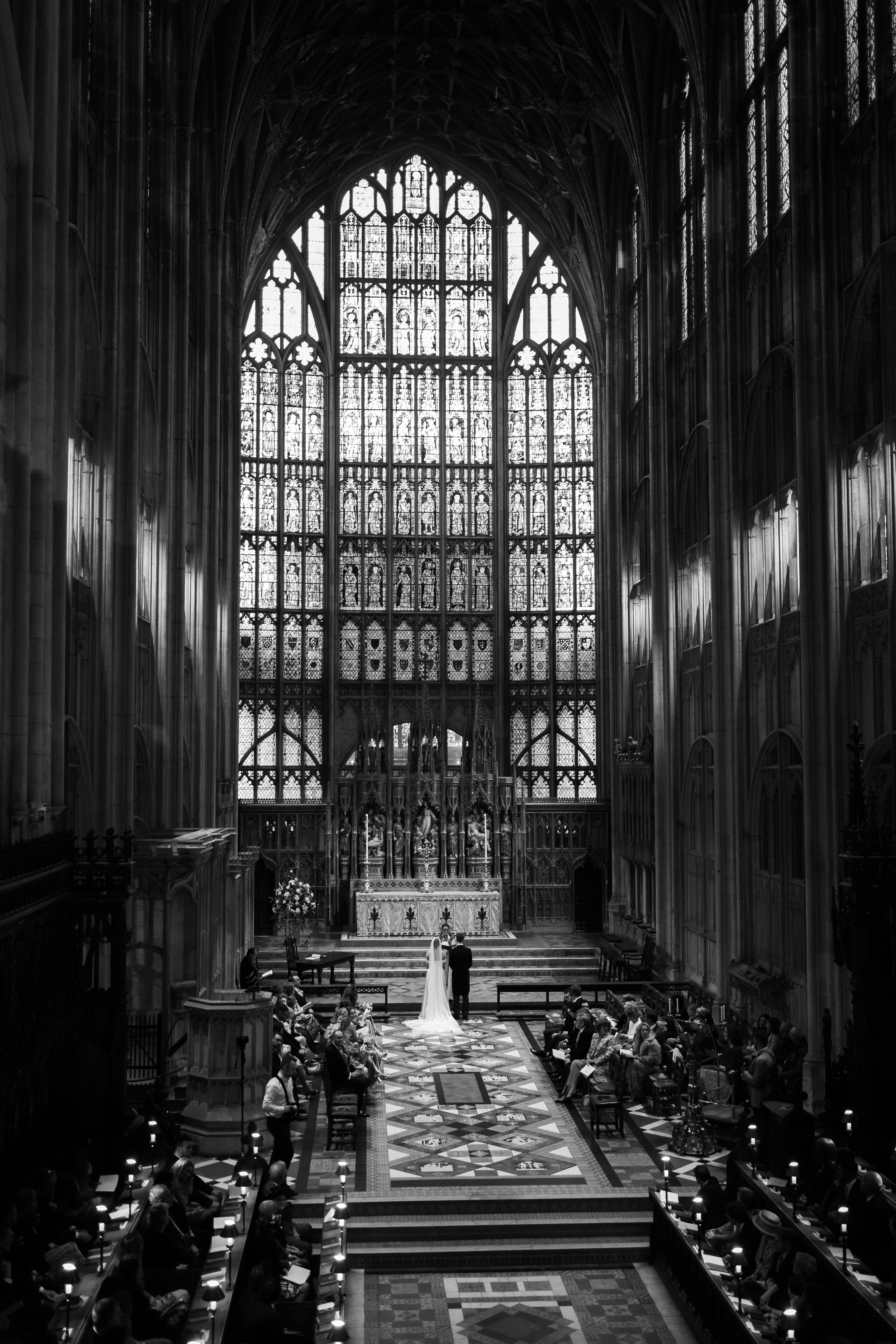 Black and white photo of a wedding ceremony at Gloucester cathedral with intricate stained glass windows and gothic architecture.