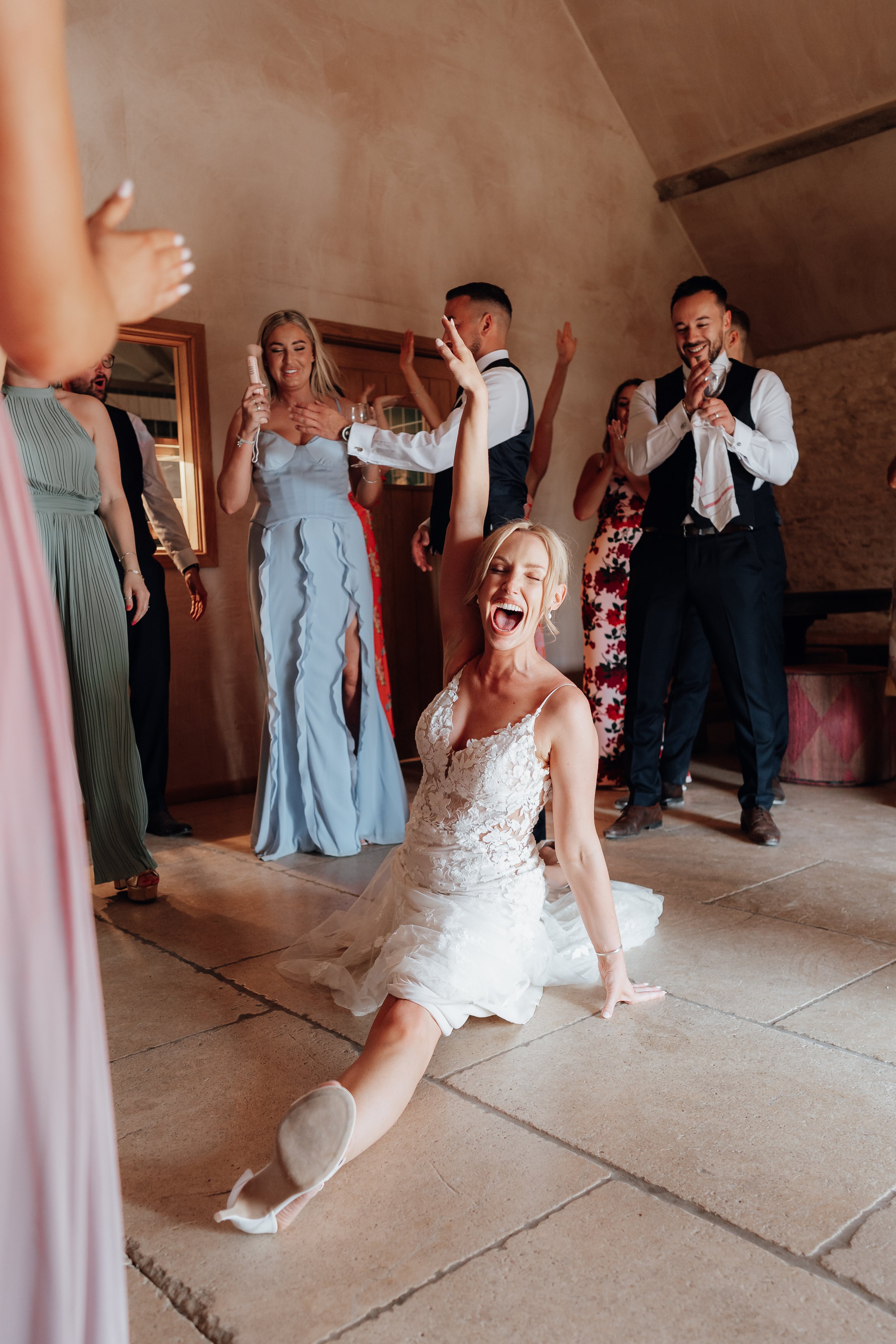Bride in lace gown doing splits with guests celebrating around her indoors
