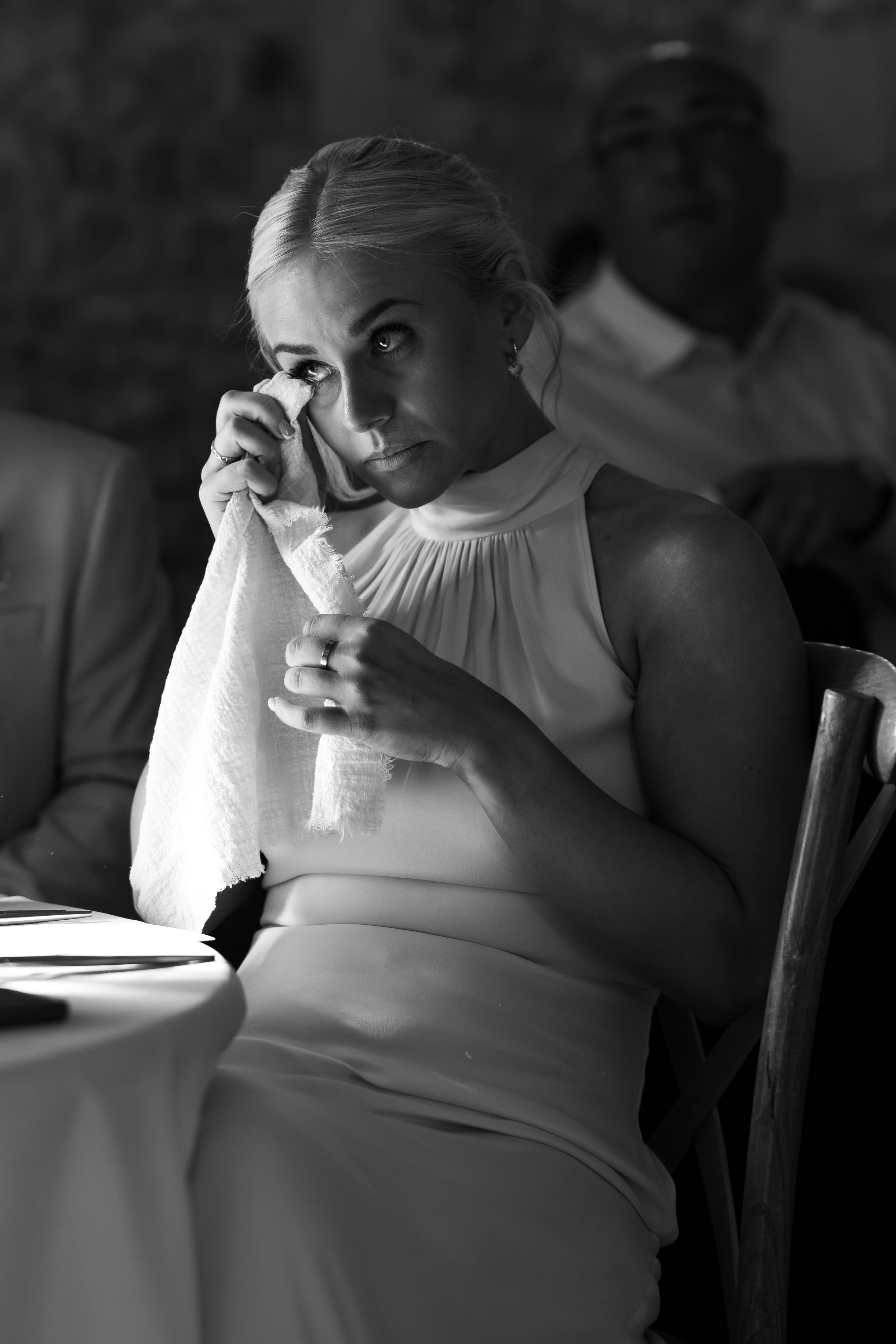 A woman in a sleeveless dress wiping her eyes with a tissue, appearing emotional, sitting at a table in a dimly lit setting.