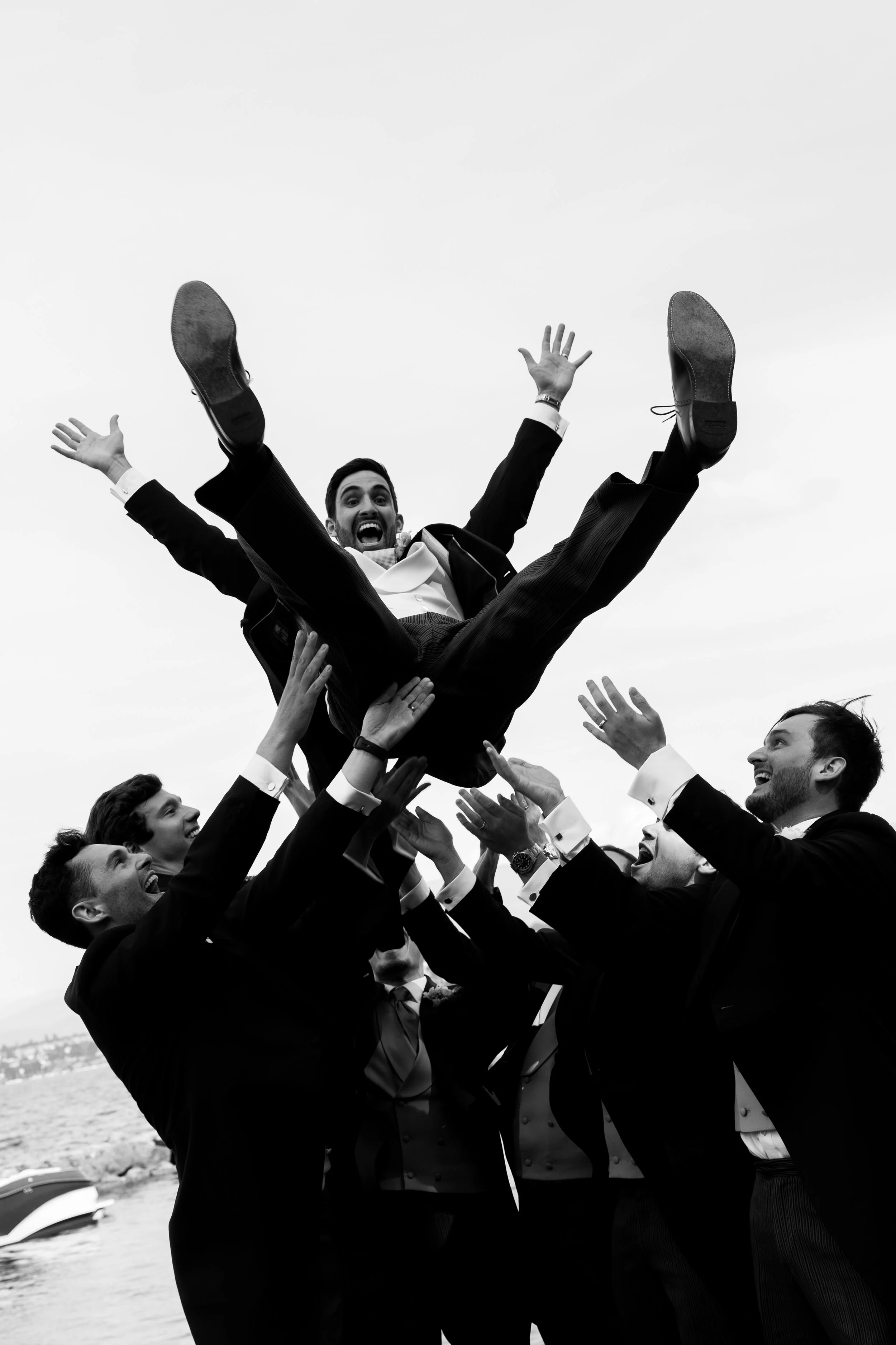 A group of men in tuxedos are lifting a man in a tuxedo into the air during a celebration near a body of water.