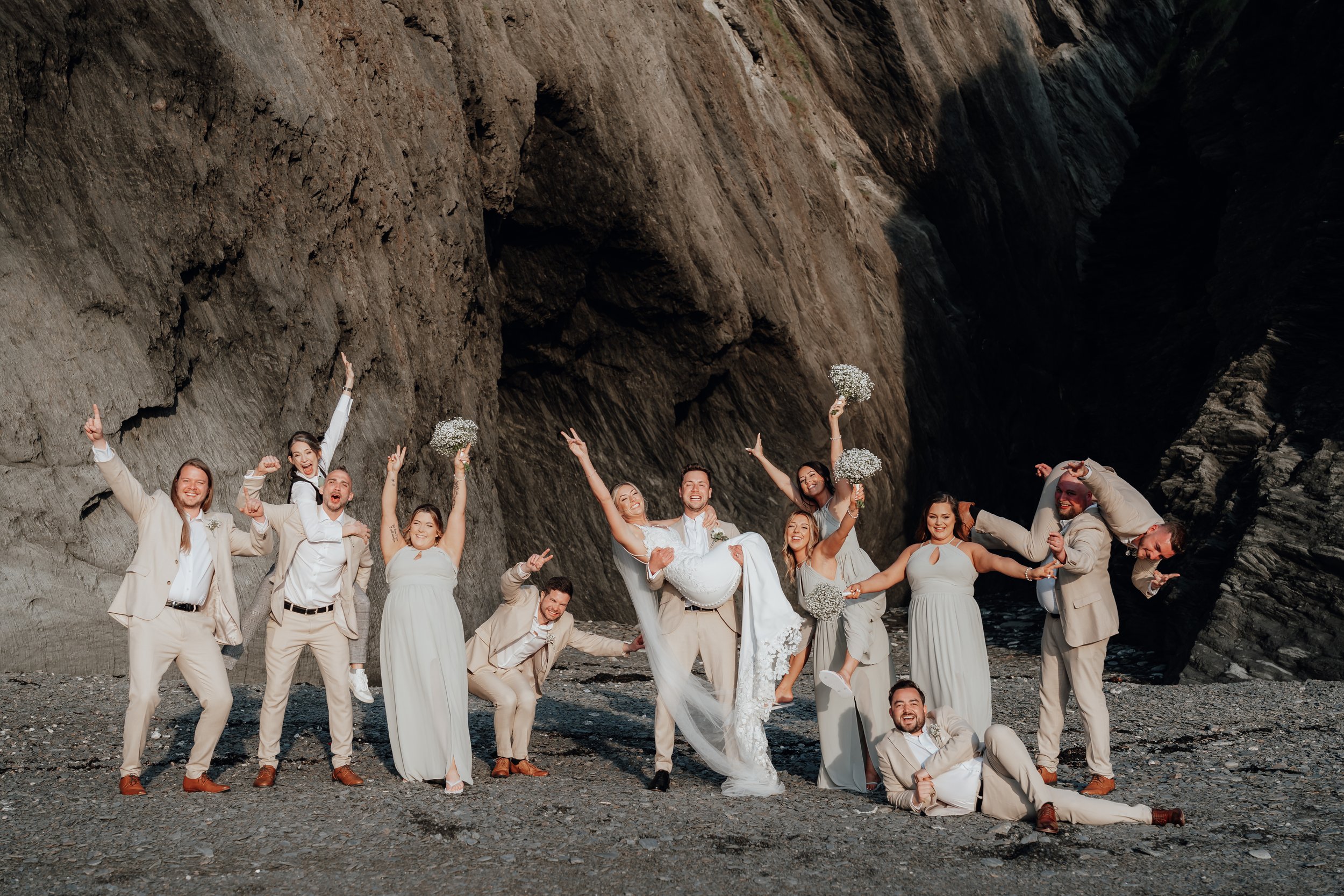 A group of people wearing formal attire, including a bride in a white dress and groom in a suit, posing playfully on a rocky beach with a cliff backdrop.
