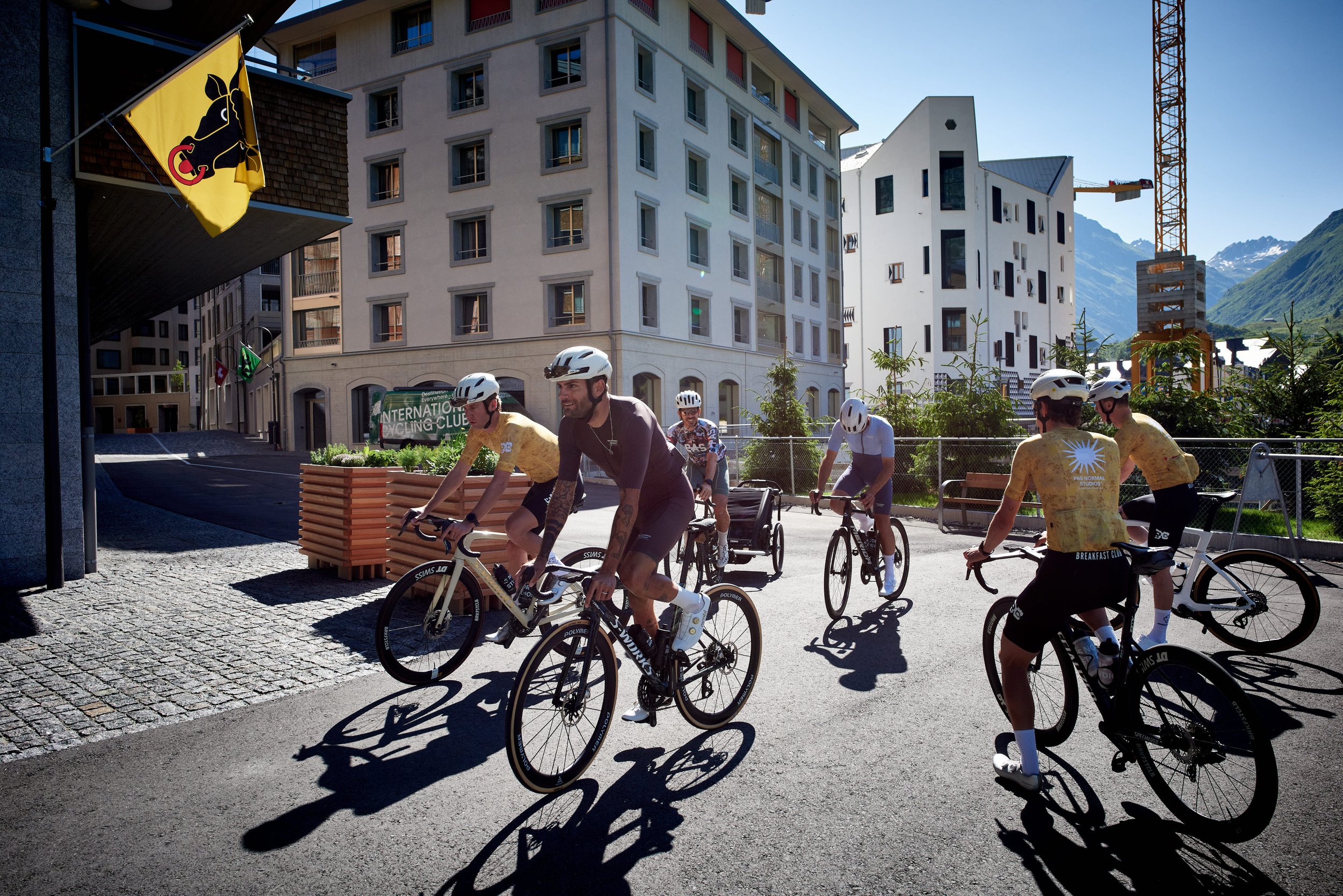 Group of cyclists riding on a city street with modern buildings, mountains in the background, and clear blue sky.