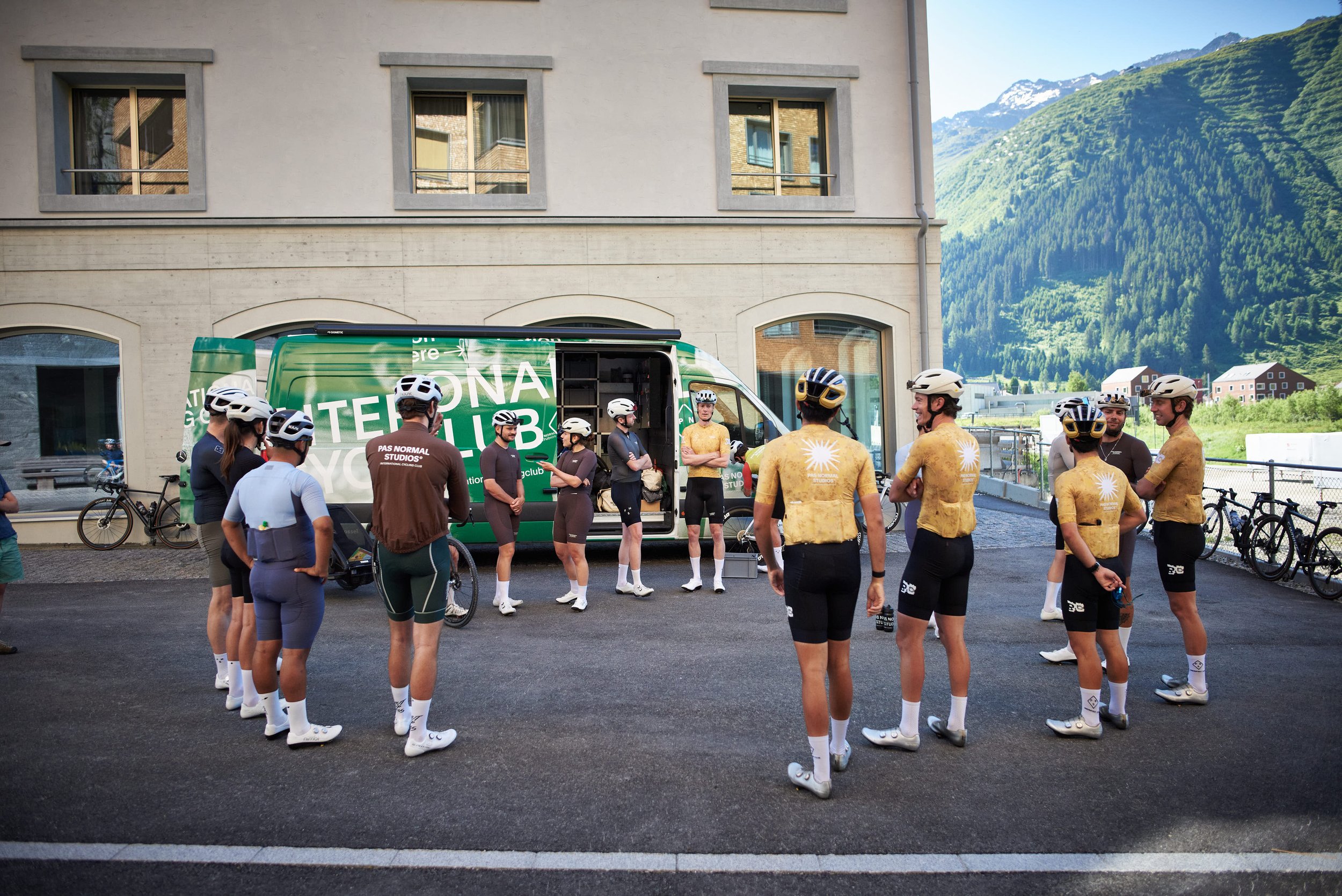 Cyclists in yellow and blue cycling outfits listening to a coach outside a green van with mountain scenery in the background.