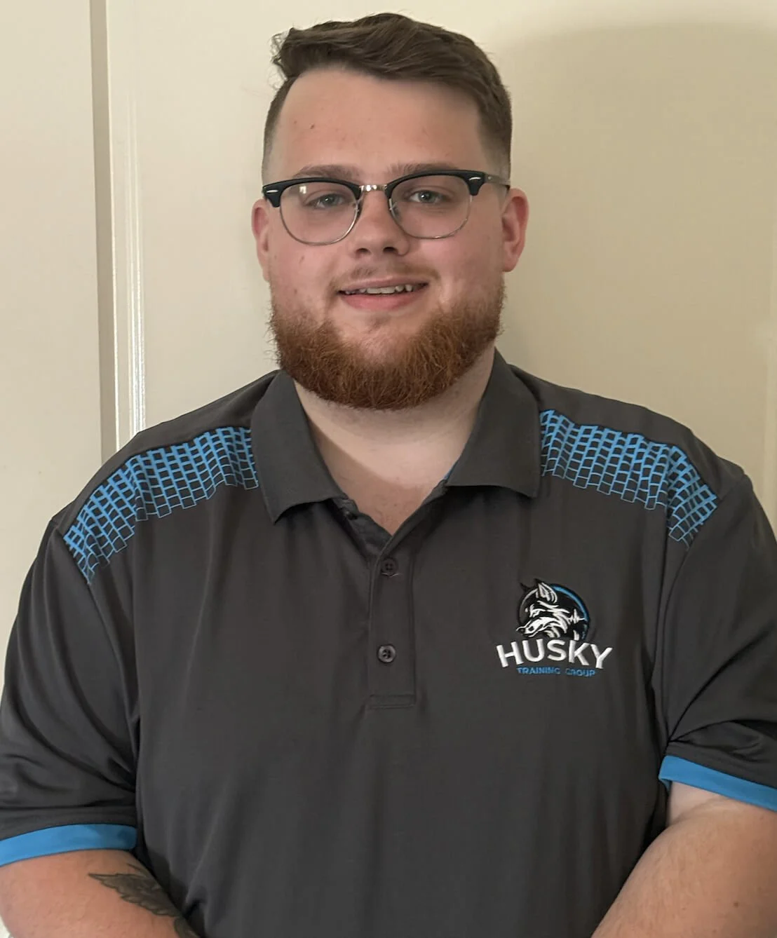A person with glasses and a beard wearing a gray shirt with the logo "Husky Training Group."