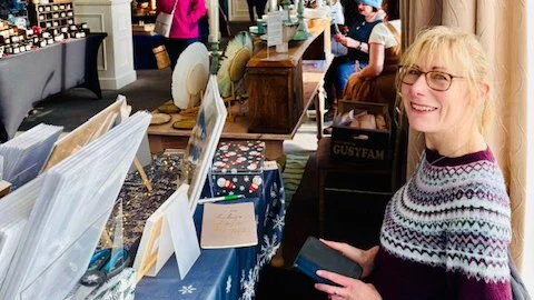 Sarah Elliott at her market stall selling her artwork, with other shoppers in the background.
