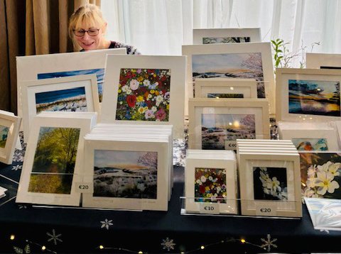 Sarah Elliott selling her framed and unframed photographs and artwork at a table, with various scenic and floral images, some with price tags.