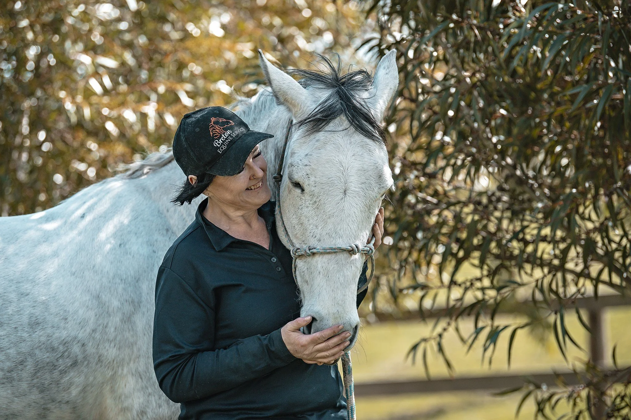 A woman in a black polo shirt and cap smiling and gently touching a white horse's face, both standing outdoors with trees and foliage in the background.