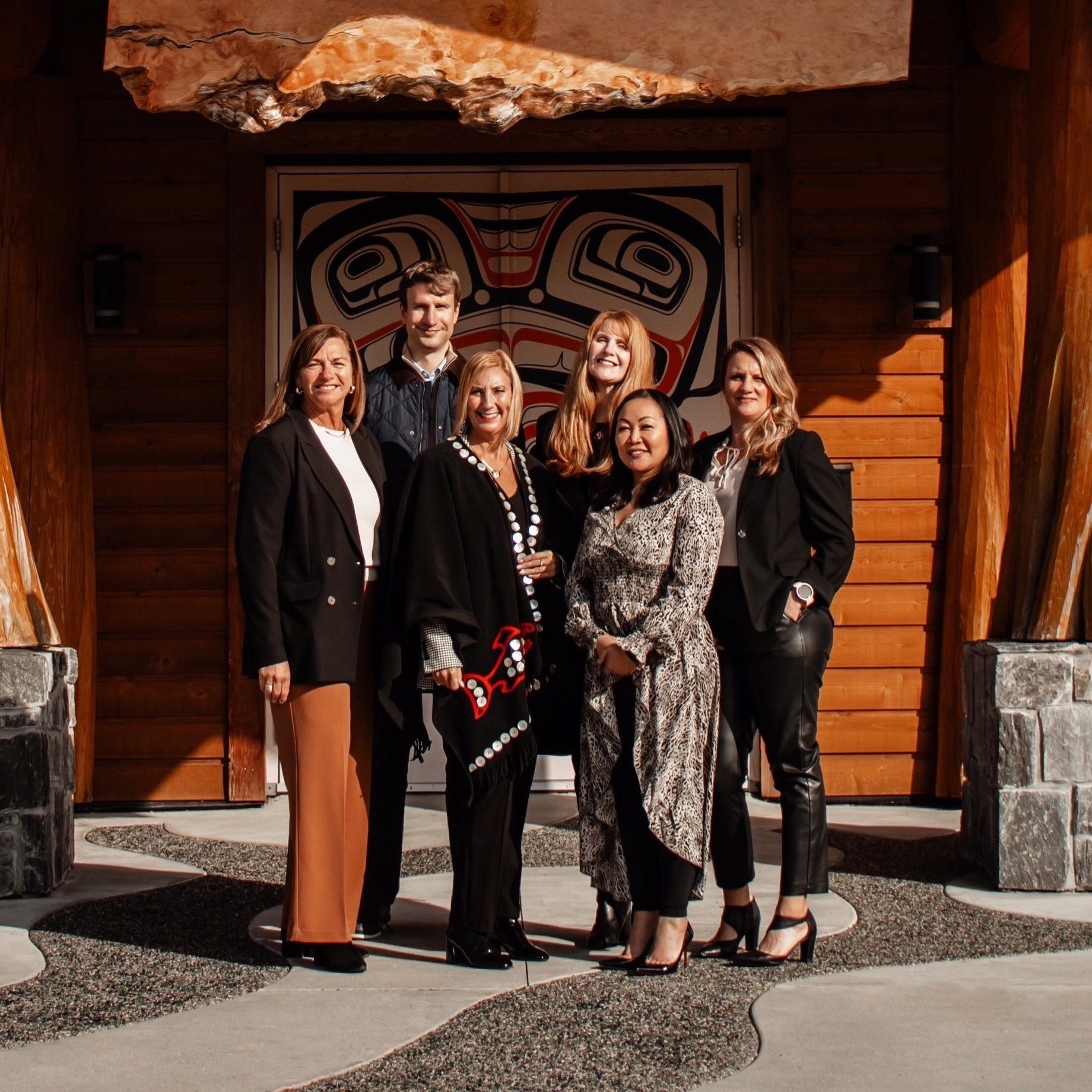 A group of seven people posing outside a building with wooden siding and a large Northwest Coast Indigenous art design on the door. They are dressed in business casual attire, smiling at the camera.