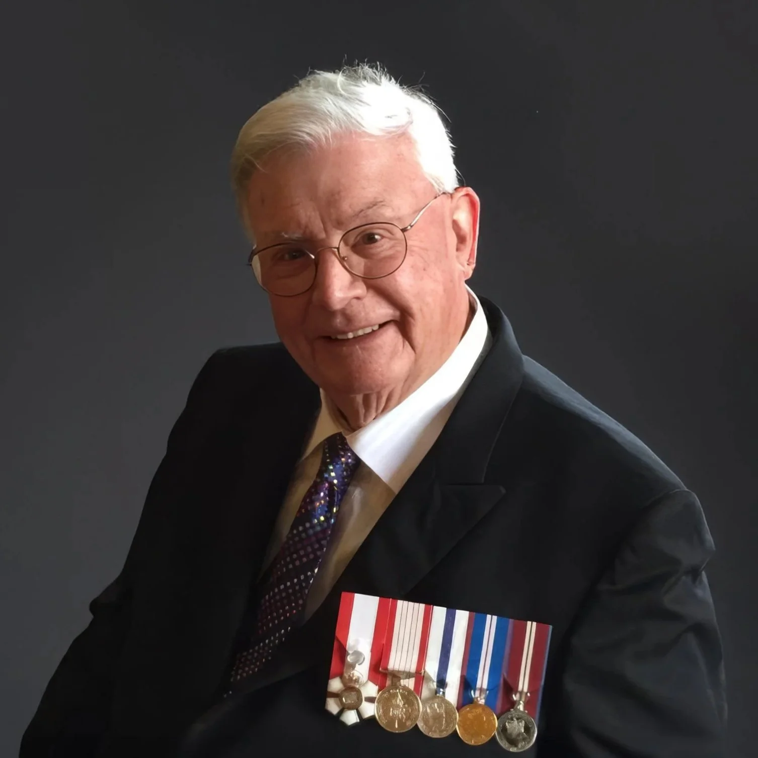 An elderly man wearing glasses and a black suit with a white shirt and a dark tie with colorful dots. He has white hair and is smiling. He is wearing several medals on his suit jacket.