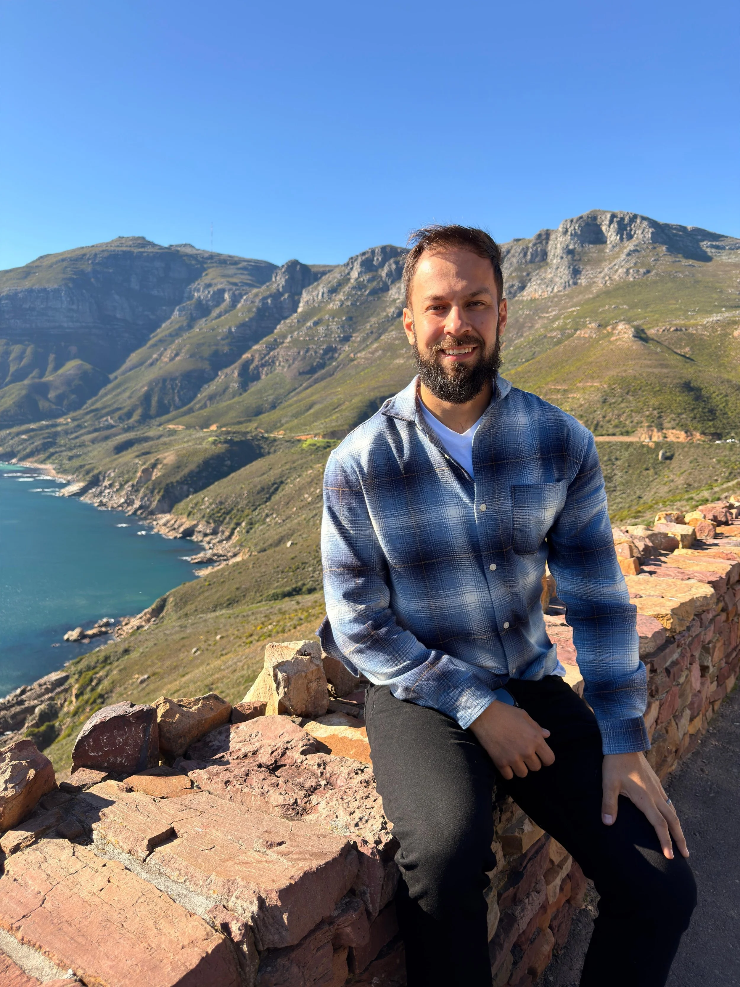 A man with a beard and short hair, wearing a plaid shirt and black pants, sitting on a stone railing with mountains, greenery, and a body of water in the background under a clear blue sky.