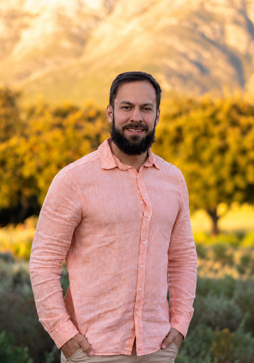 A man with dark hair and a beard smiling outdoors during autumn, wearing a light pink button-up shirt, standing in front of a background of trees with yellow leaves and mountains.