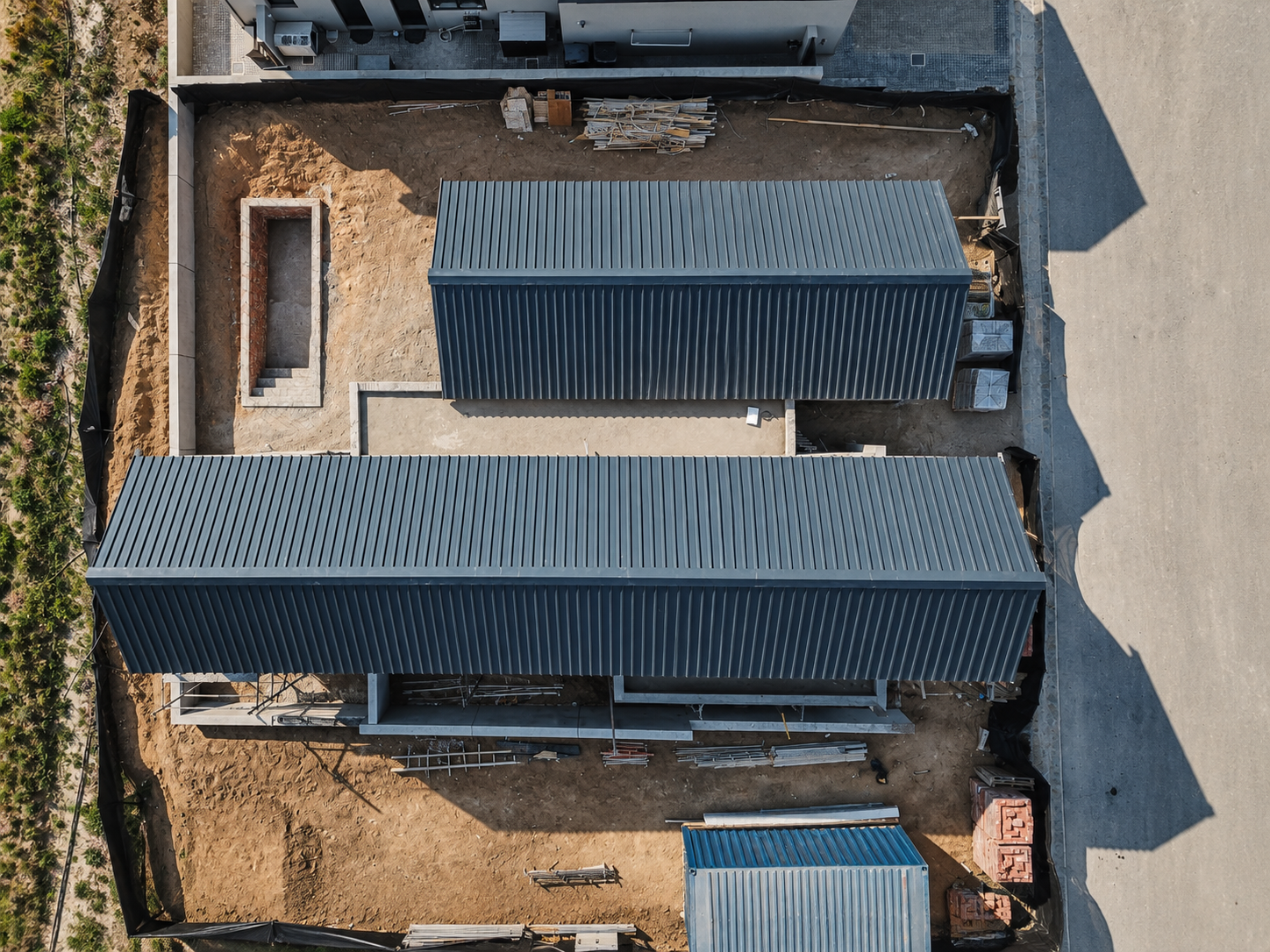An aerial view of a construction site showing two metal-roofed structures, a rectangular pool-shaped foundation, construction materials, and a sidewalk.