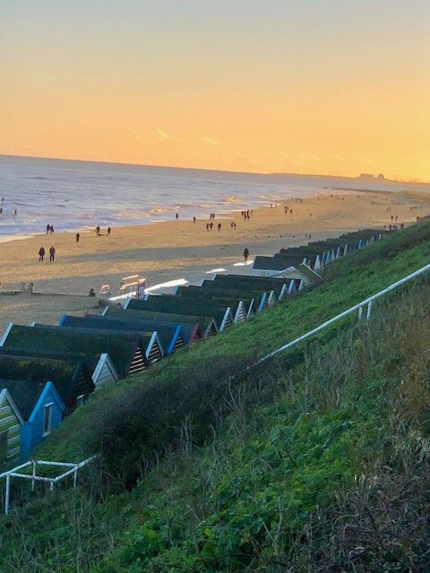 Beach with colorful beach huts, people walking, and a sunset sky over the ocean.