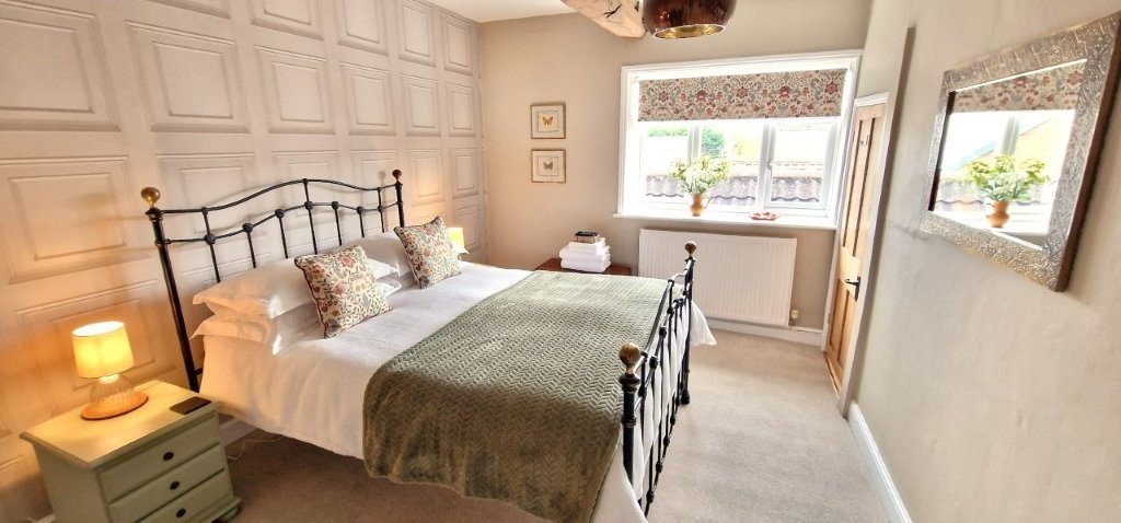 A cozy bedroom with a large bed featuring white linens, decorative pillows, and a green textured bedspread. There's a bedside table with a small lamp, a window with a floral valance, wall art, and a mirror with a glittery frame. The room has neutral